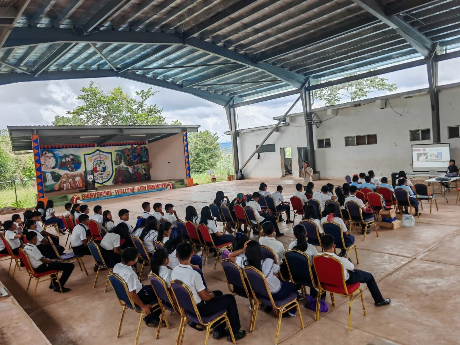 Niños en uniforme escolar sentados en sillas en un auditorio con estructura metálica, asistiendo a una presentación con una pantalla y una mujer hablando.