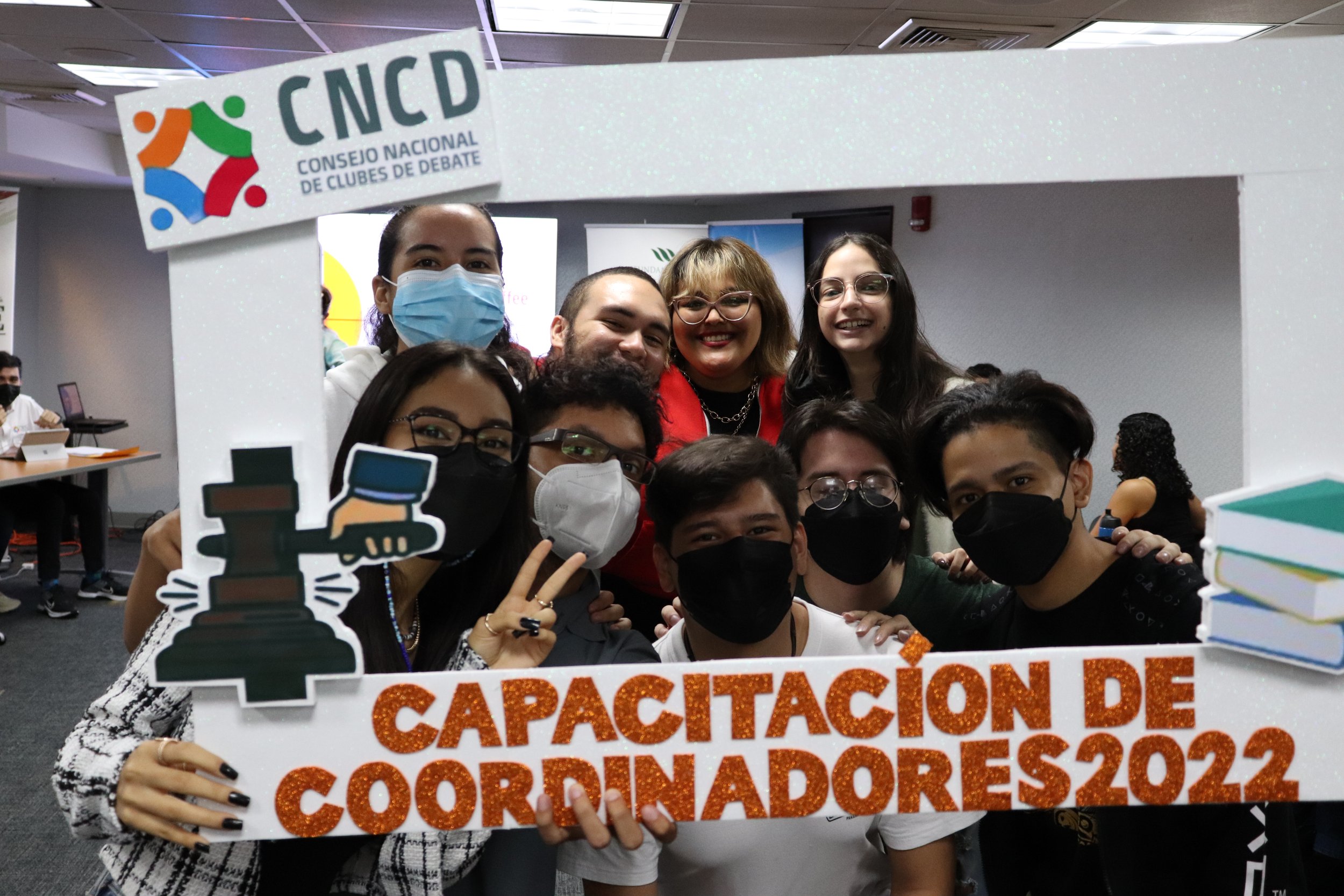 Grupo de personas con mascarillas celebrando en un evento, sosteniendo un cartel que dice 'Capacitación de Coordinadores 2022' y un cartel del Consejo Nacional de Clubes de Debate.