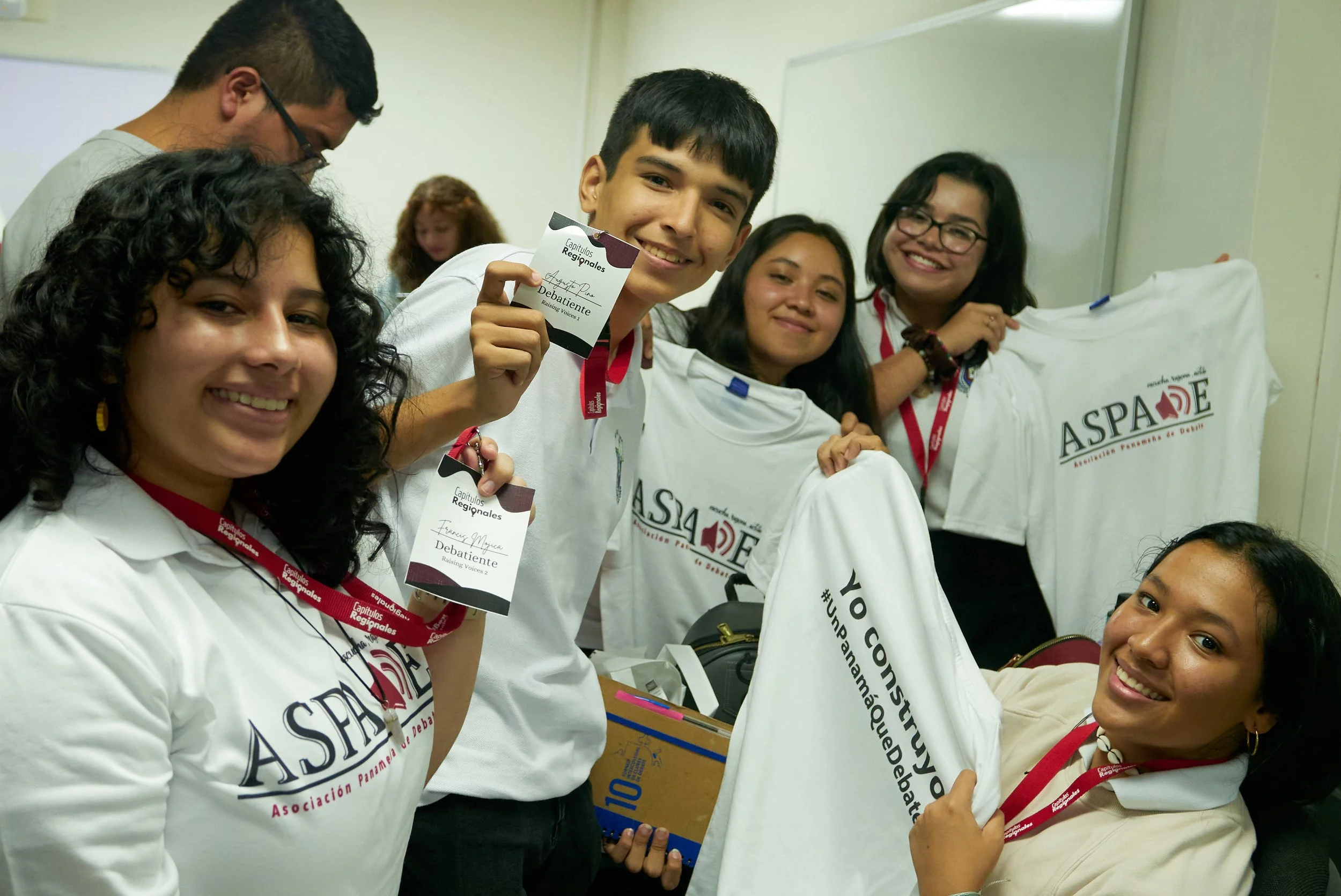 Grupo de jóvenes en un aula enfocada en un joven que muestra una tarjeta que dice 'Capítulos Regionales', todos llevan camisetas blancas con el logotipo de ASPA ¿de debate. Algunos sonríen y usan gafas.