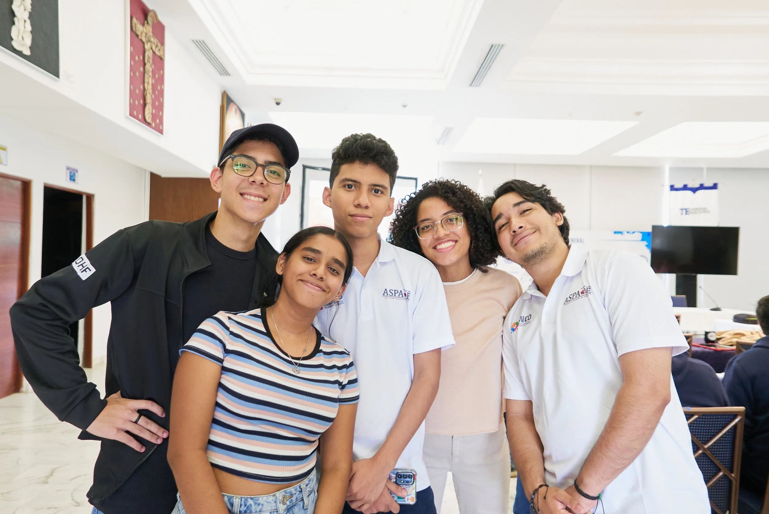 Grupo de cinco jóvenes sonriendo en un espacio interior, algunos con gafas y ropa casual, otros con camisetas blancas con logotipos y letras, en un ambiente bien iluminado con paredes blancas y decoración en el fondo.