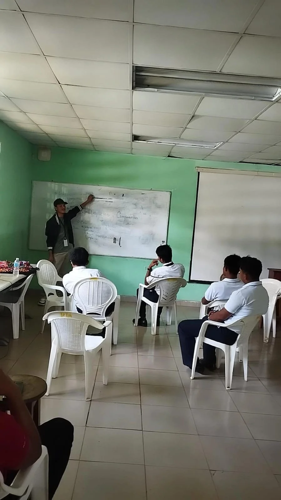 Un grupo de estudiantes mira a un profesor que enseña en una pizarra en un aula escolar, con sillas y mesas blancas en un entorno de paredes verdes y techo de baldosas.