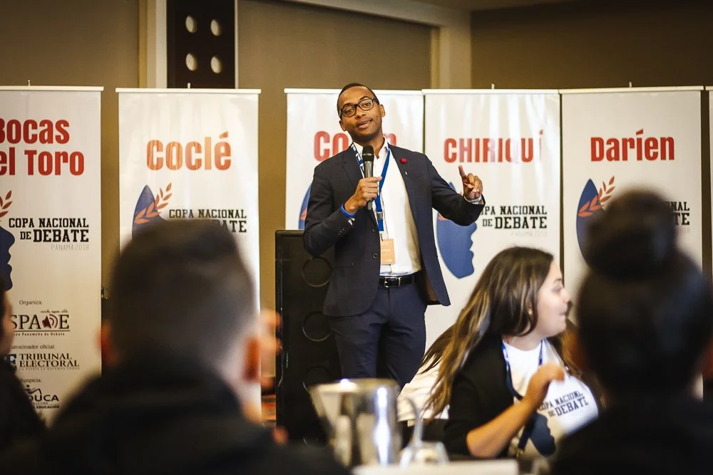 Un hombre con gafas de vestir y saco oscuro da un discurso con micrófono en un evento de debate, con varias pancartas en el fondo que muestran palabras como 'bocas del toro', 'coclè', 'chiricu', y 'darien', en un espacio de conferencia.