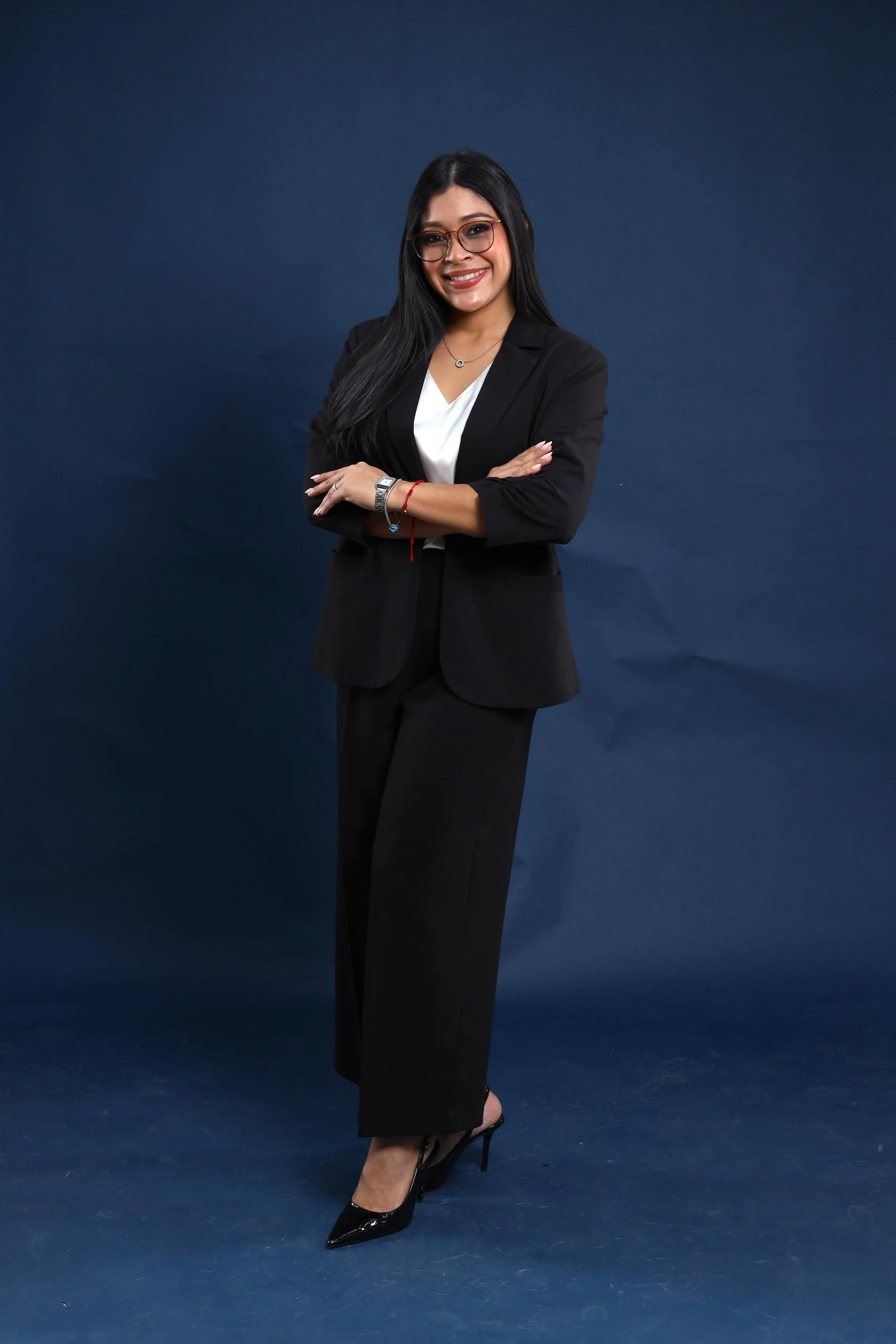 Mujer sonriendo con gafas y cabello largo, vestida con traje negro y zapatos de tacón, posando con los brazos cruzados frente a un fondo azul oscuro.