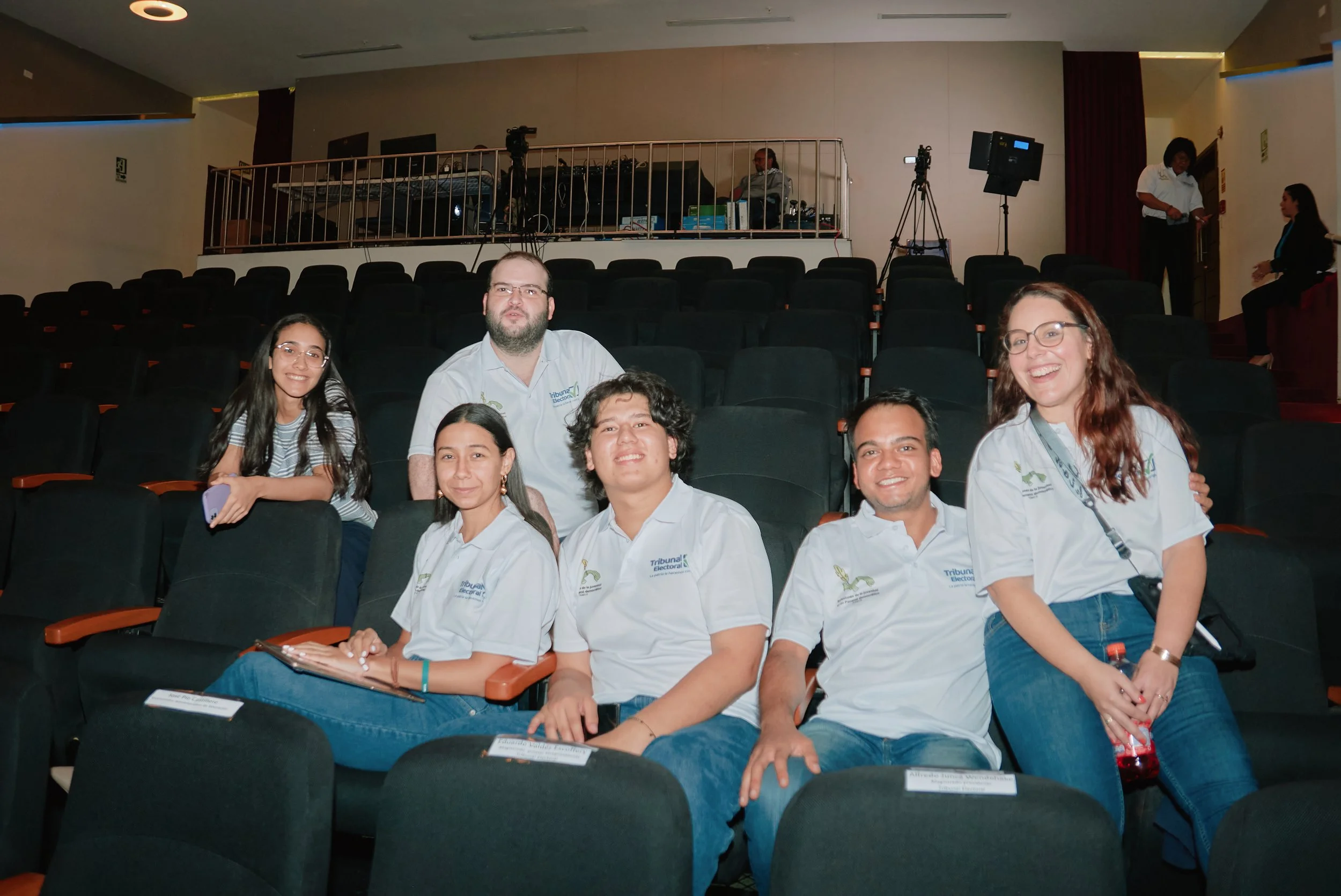 Grupo de personas sentadas en un auditorio, con equipo de sonido y cámaras en el fondo, parecen participar en un evento o conferencia.