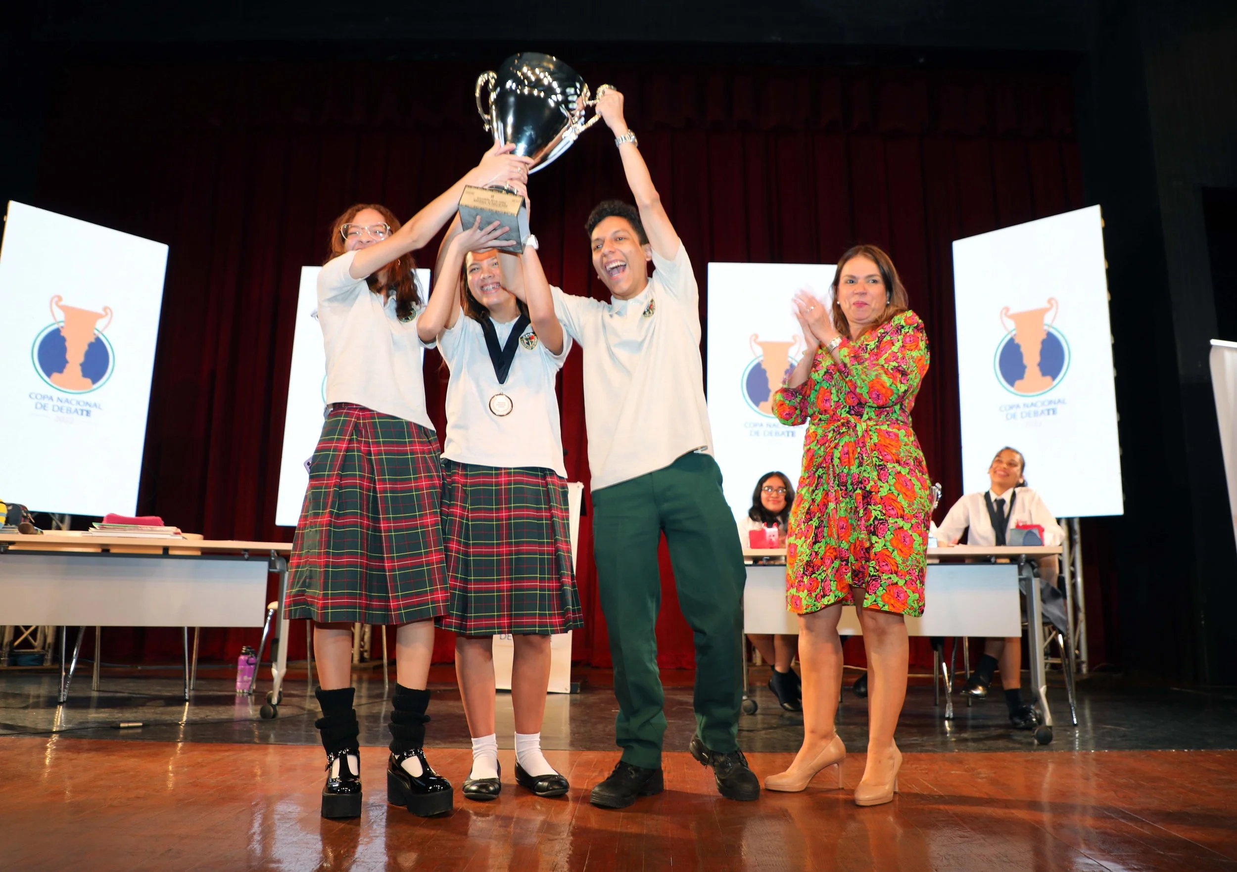 Jóvenes en uniforme escolar celebrando con un trofeo en un evento de debate, con una mujer en vestido colorido aplaudiendo en un escenario con pantallas que dicen 'Copa Nacional de Debate'.