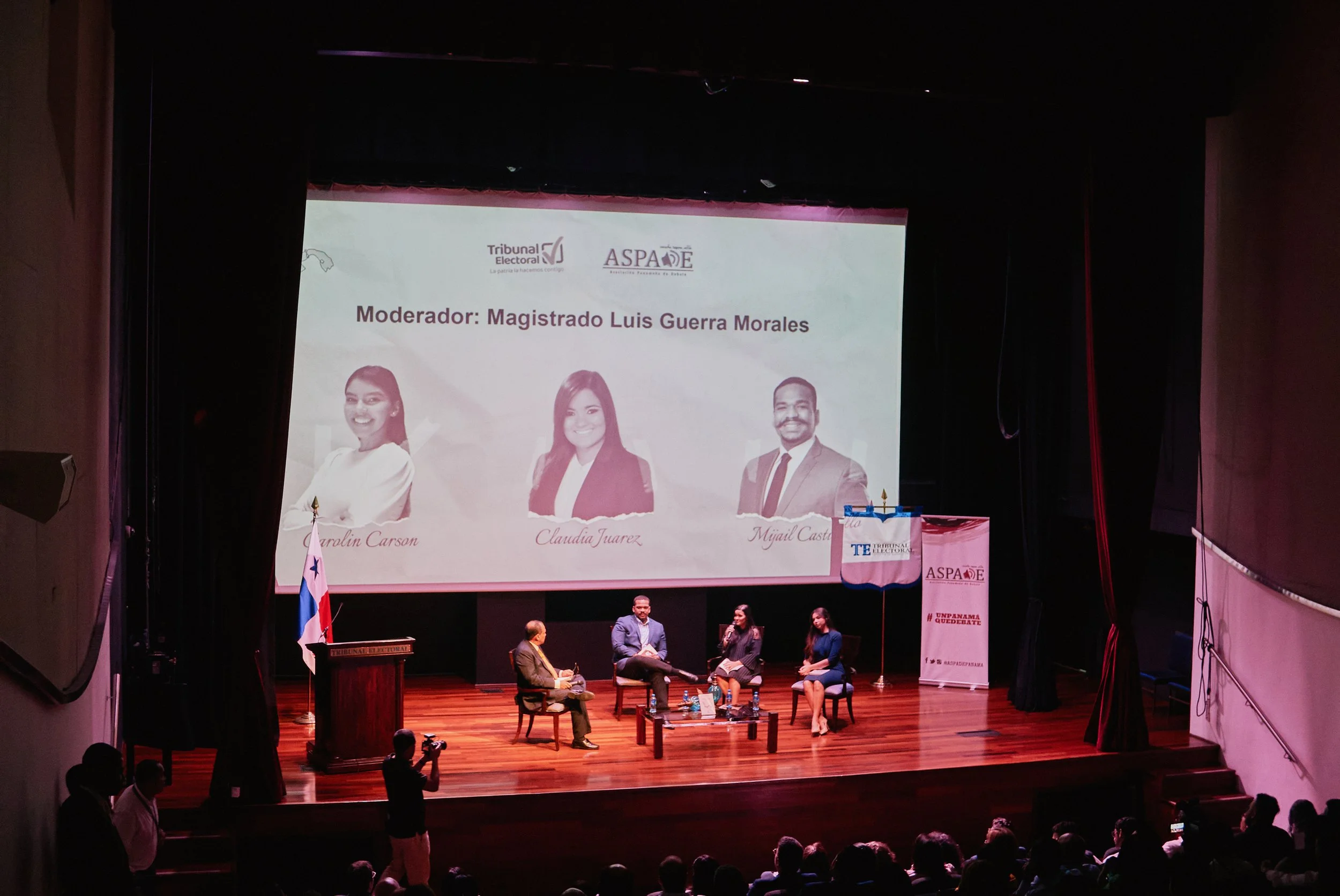 Escenario de un evento con pantalla grande, personas sentadas en el escenario y en el público, banderas, y una conferencia sobre justicia electoral en un teatro.