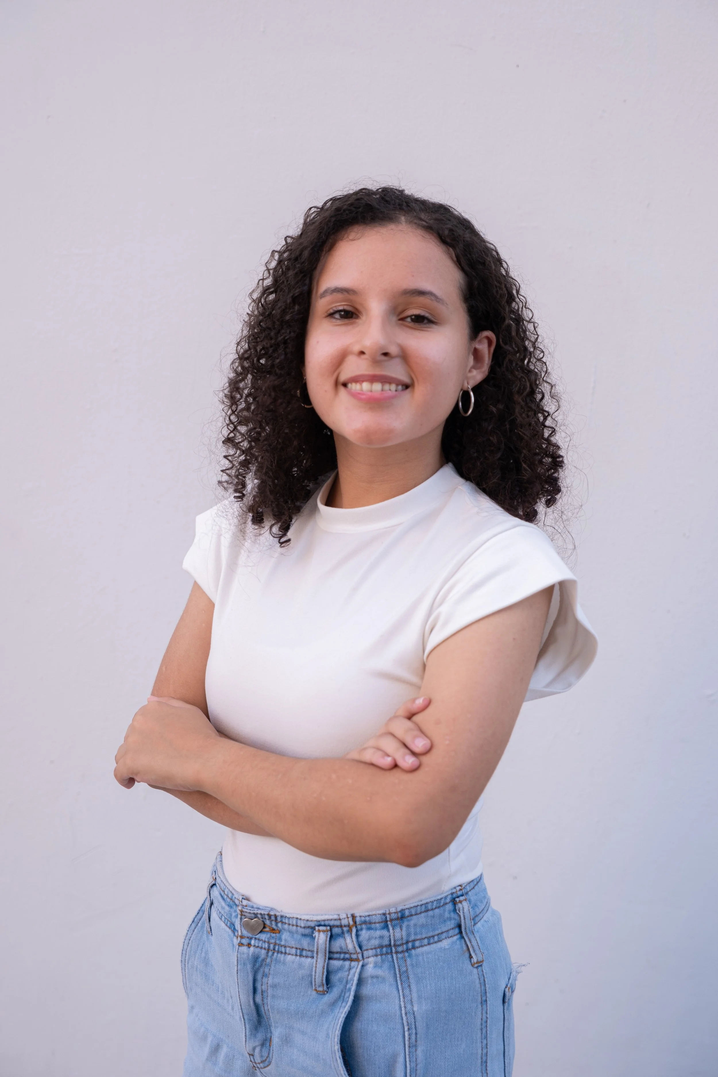 Retrato de una joven con cabello rizado, de complexión media, vistiendo una camiseta blanca y pantalones vaqueros, sonriendo y con los brazos cruzados frente a un fondo gris.