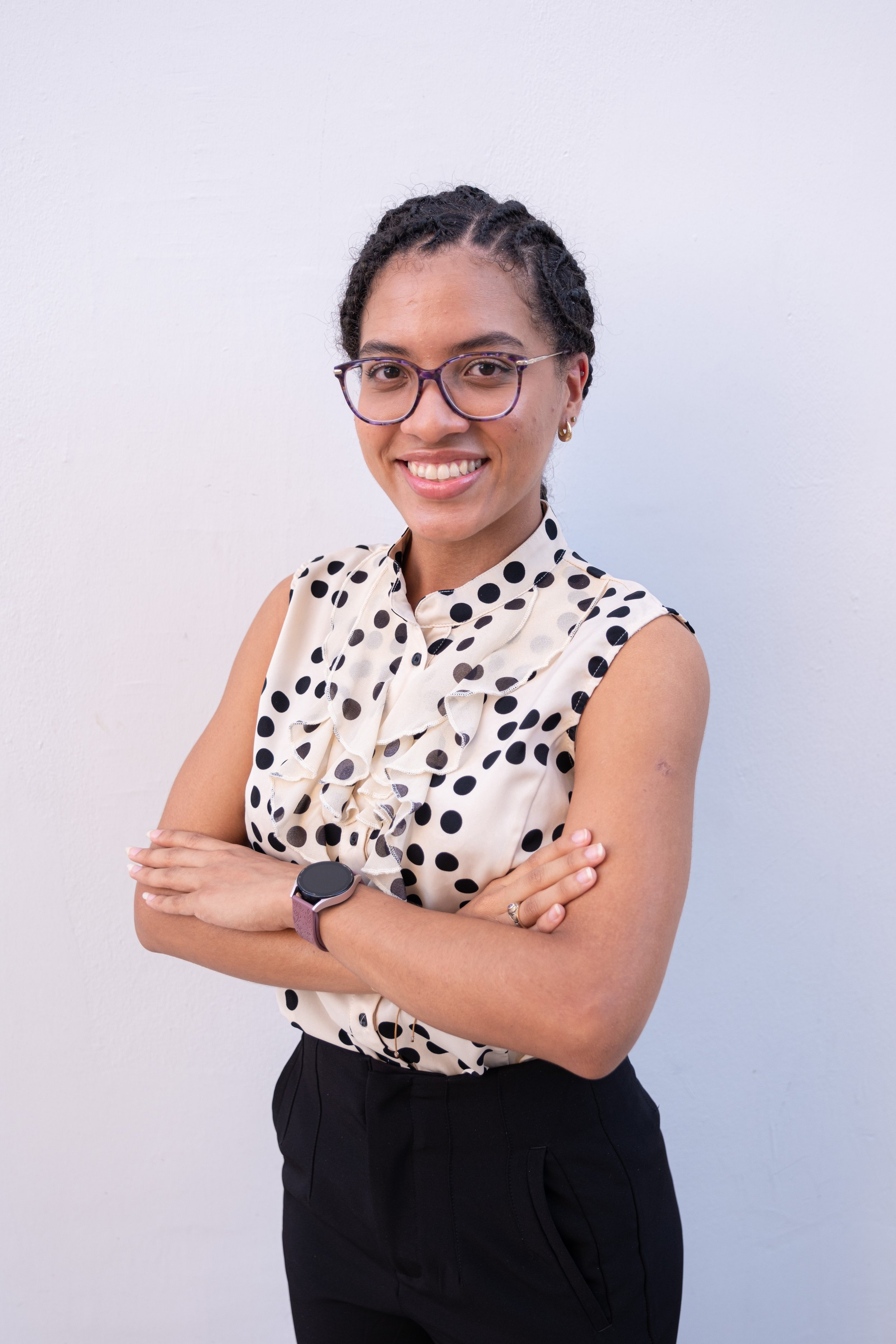 Mujer joven con gafas y traje de polka, sonriendo con los brazos cruzados frente a una pared blanca.