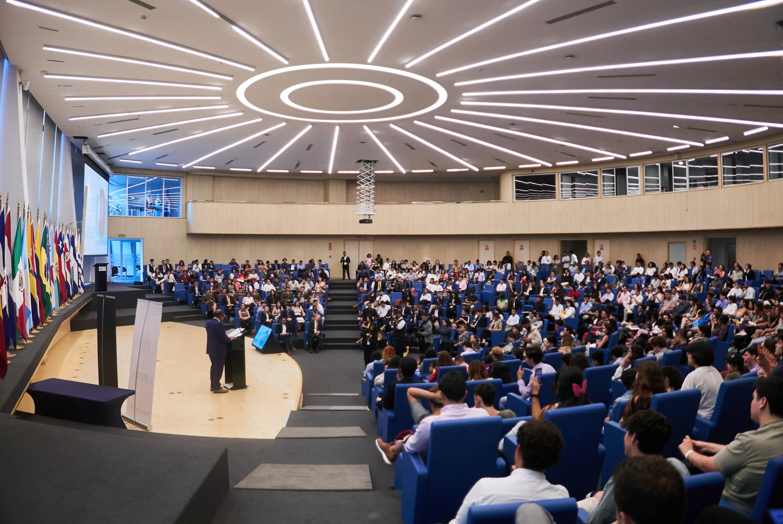 Una conferencia en un auditorio con muchas personas sentadas, un orador en el escenario y banderas de diferentes países a la izquierda del escenario.