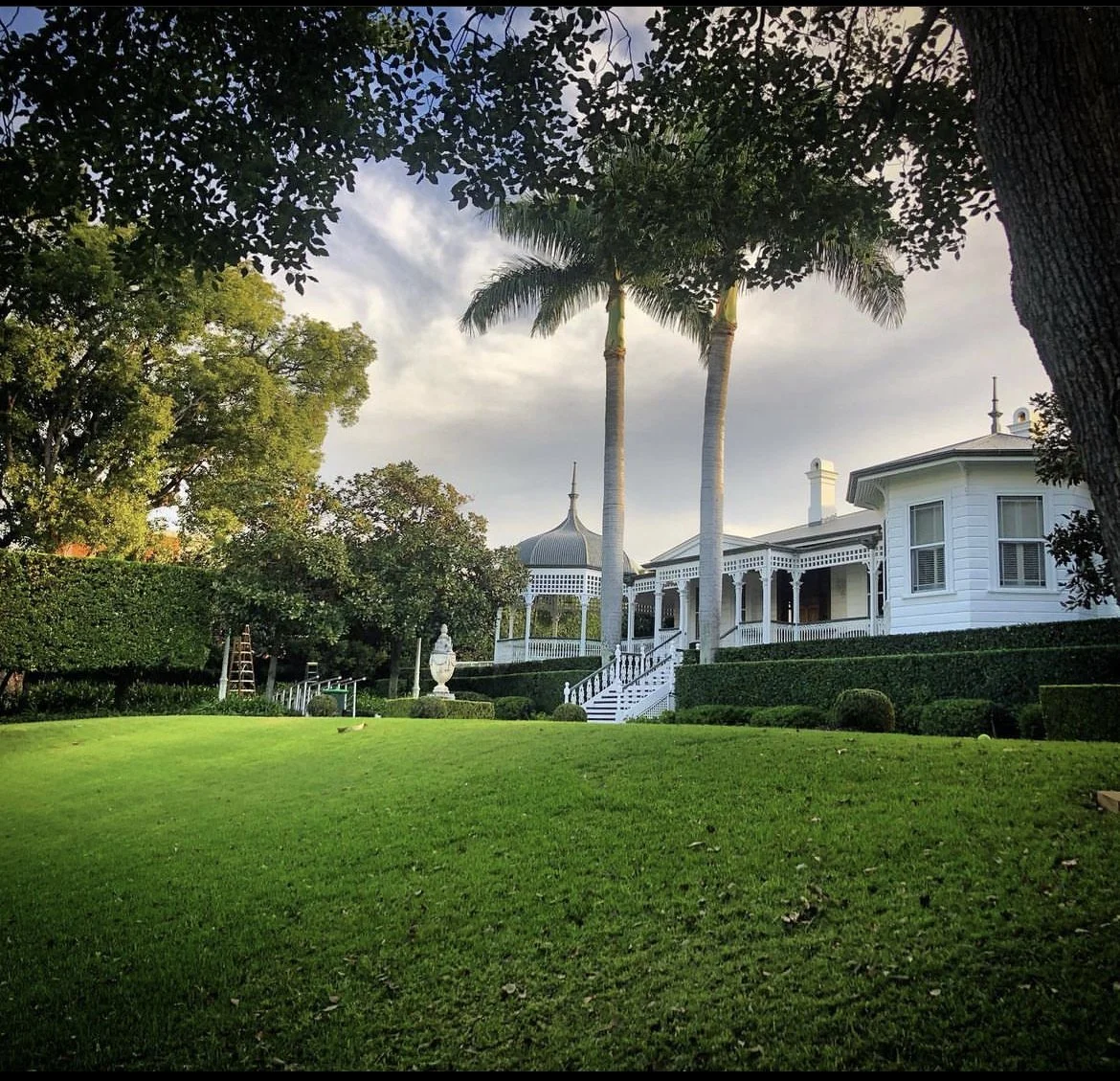 A lush green lawn in front of a white Victorian-style house with a wraparound porch, surrounded by trees including two tall palm trees, with a partly cloudy sky overhead.