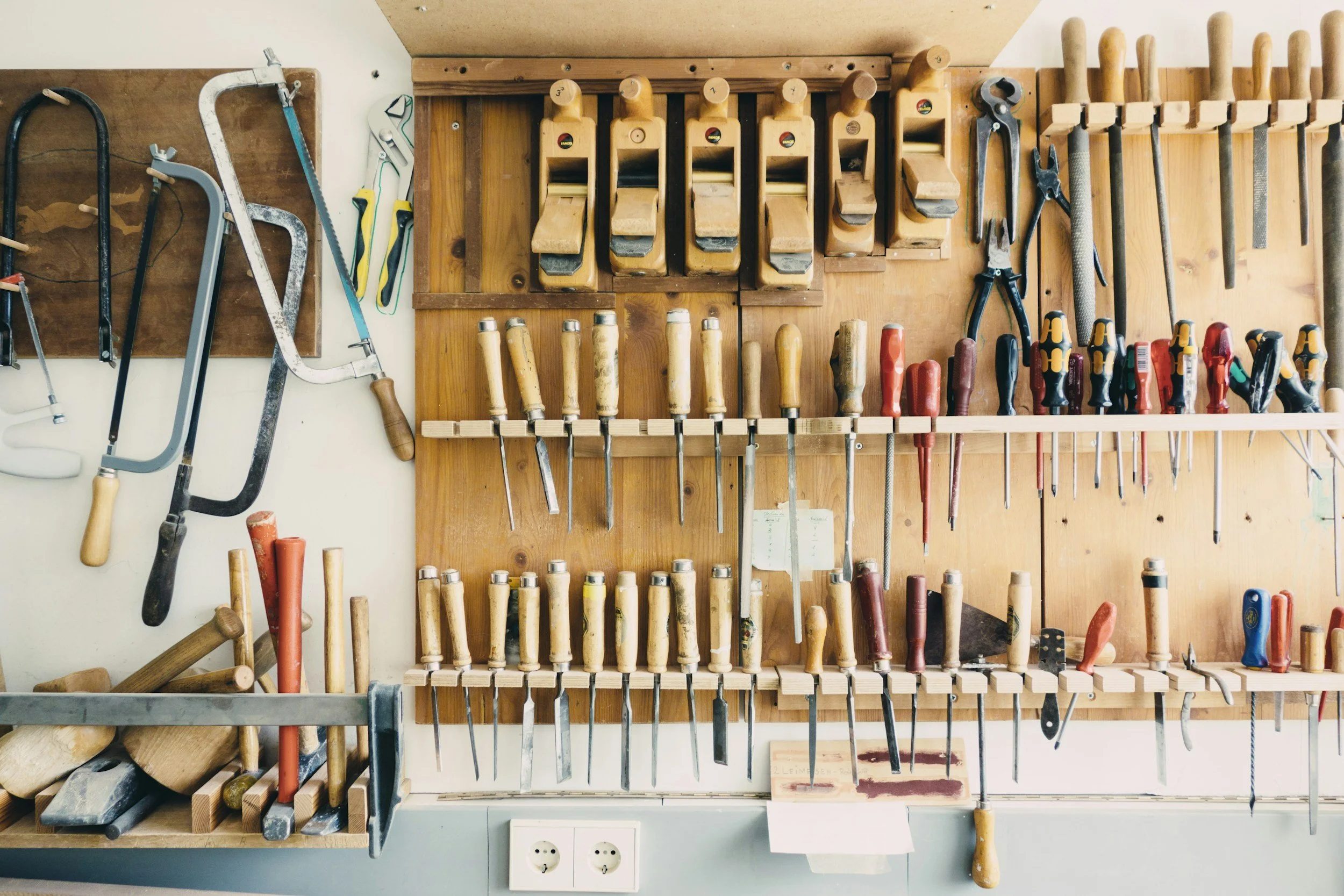 Wall-mounted woodworking tools including saws, chisels, screwdrivers, clamps, and planes in a workshop.
