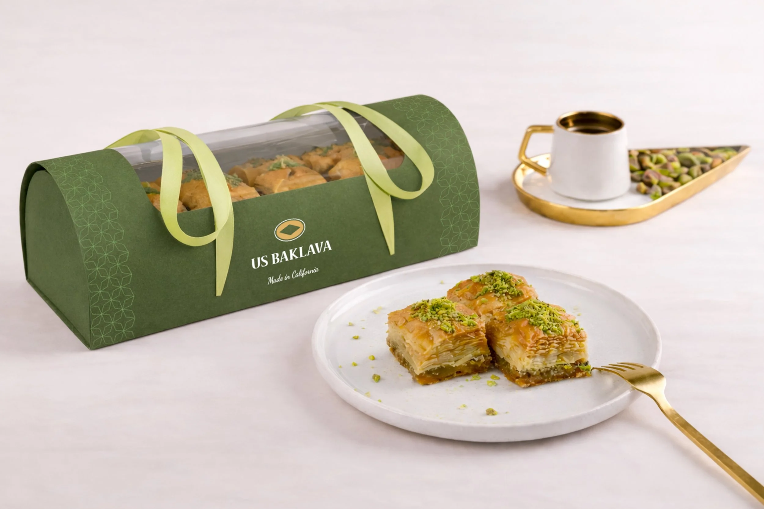 Packaged Turkish baklava with a piece on a white plate garnished with chopped pistachios, and a coffee cup with seeds on a gold tray in the background.