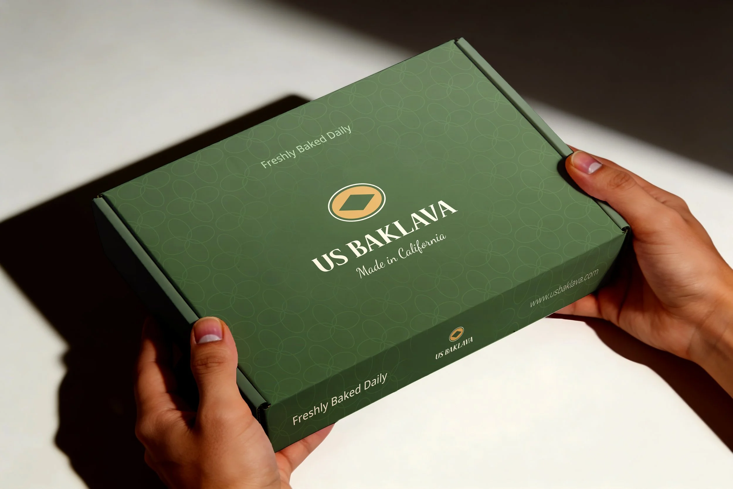 Person holding a green box of US Baklava with text indicating it's freshly baked daily and made in California.