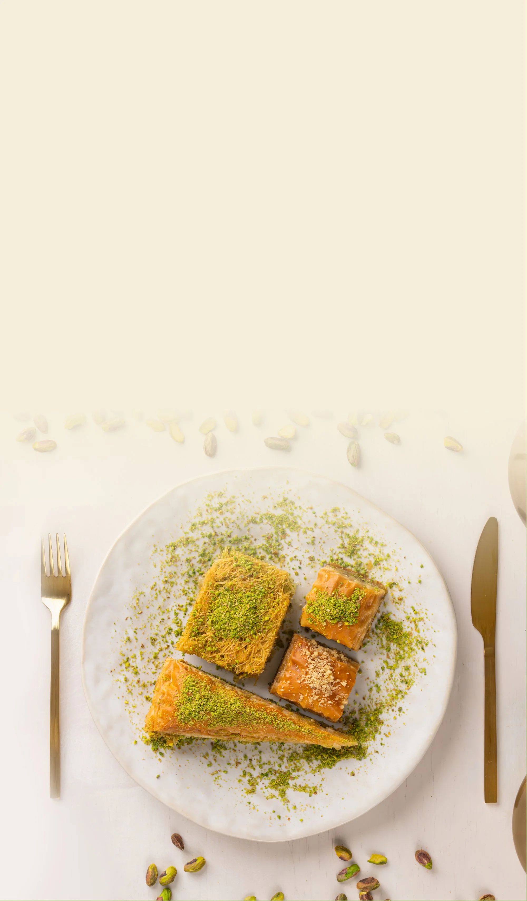 A white plate with assorted Middle Eastern desserts topped with pistachio crumbs, surrounded by scattered pistachios, a gold fork on the left, and a gold knife on the right on a white tablecloth.