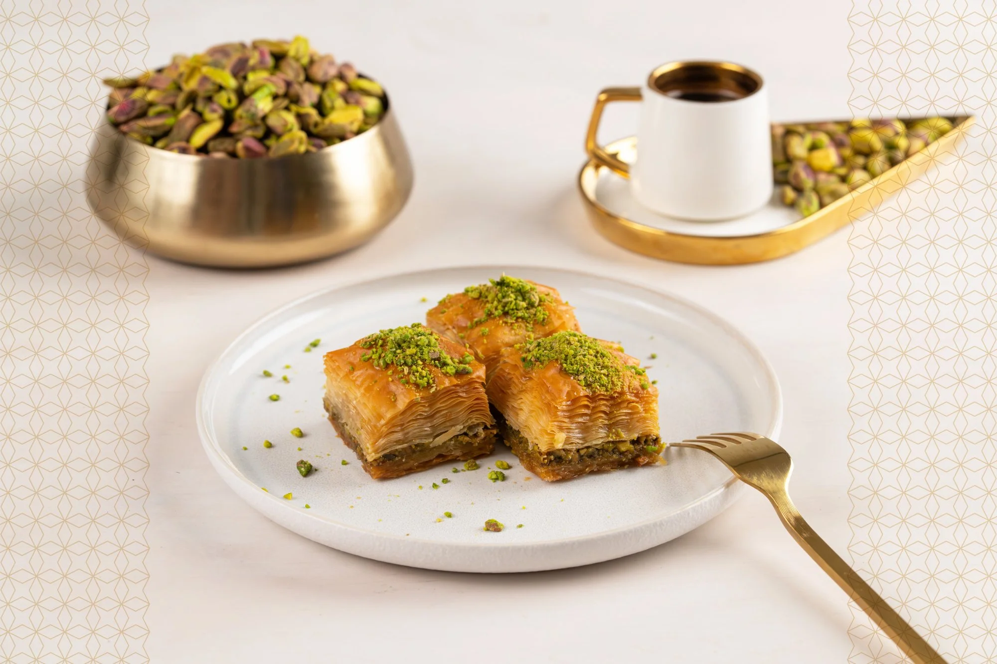 Three pieces of baklava garnished with chopped pistachios on a white plate, with a fork on the side. In the background, there is a bowl of pistachios and a cup of coffee with a saucer, all on a white table.