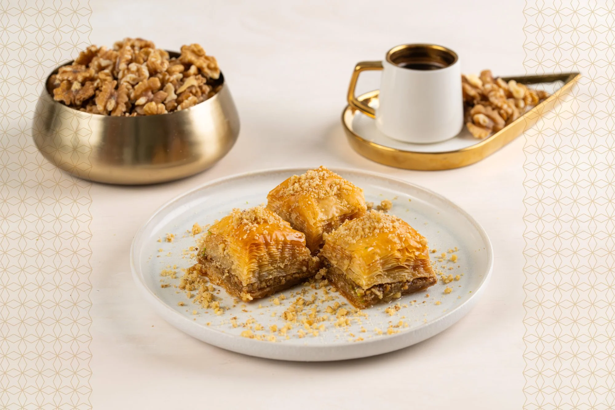 A plate of three pieces of baklava with honey and crushed nuts, a bowl of walnuts, and a cup of black coffee on a golden tray with a cup on it, all on a white table.