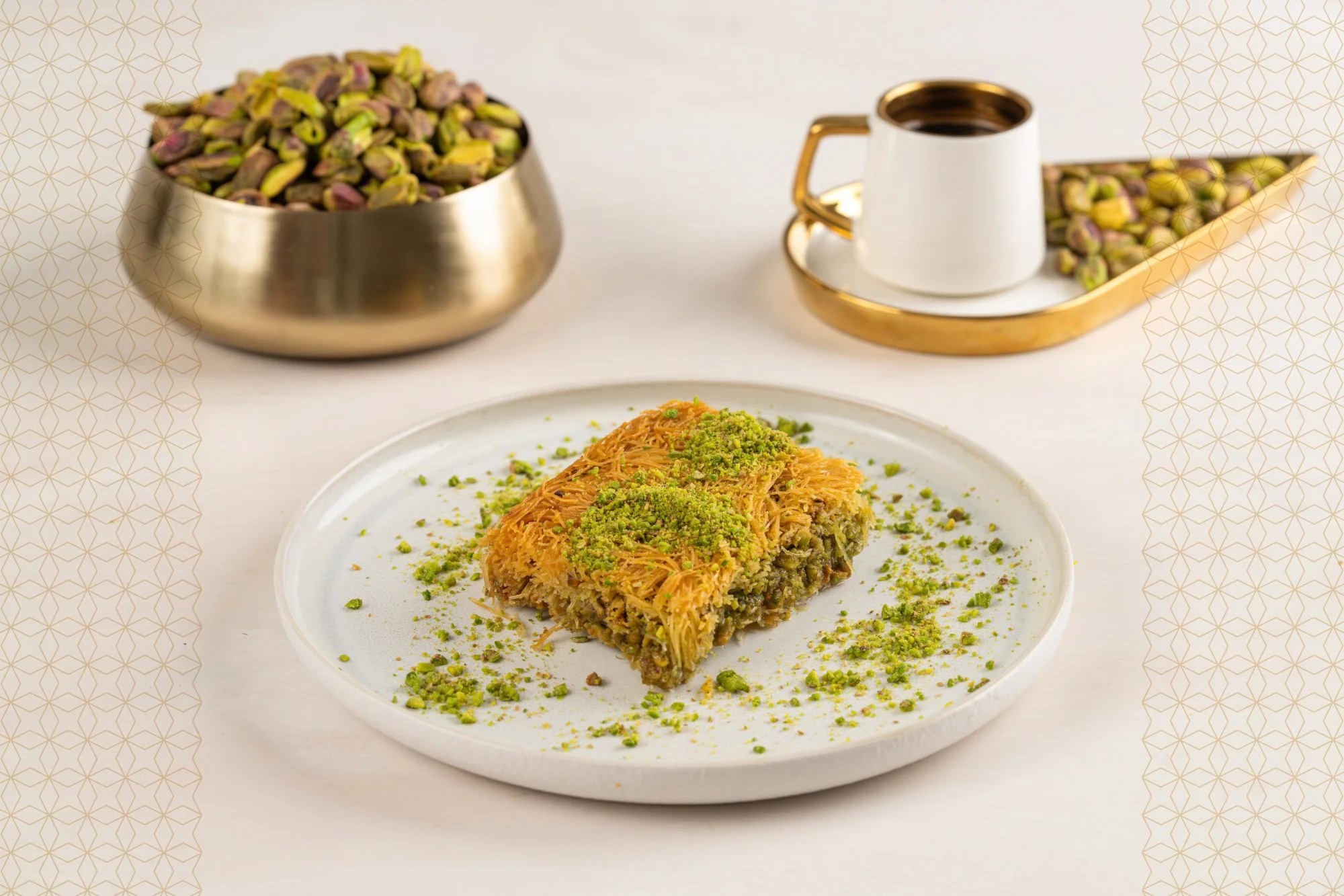 A plate of baklava garnished with chopped pistachios, with a bowl of pistachios, a cup of Turkish coffee, and a tray of pistachios on a white background.