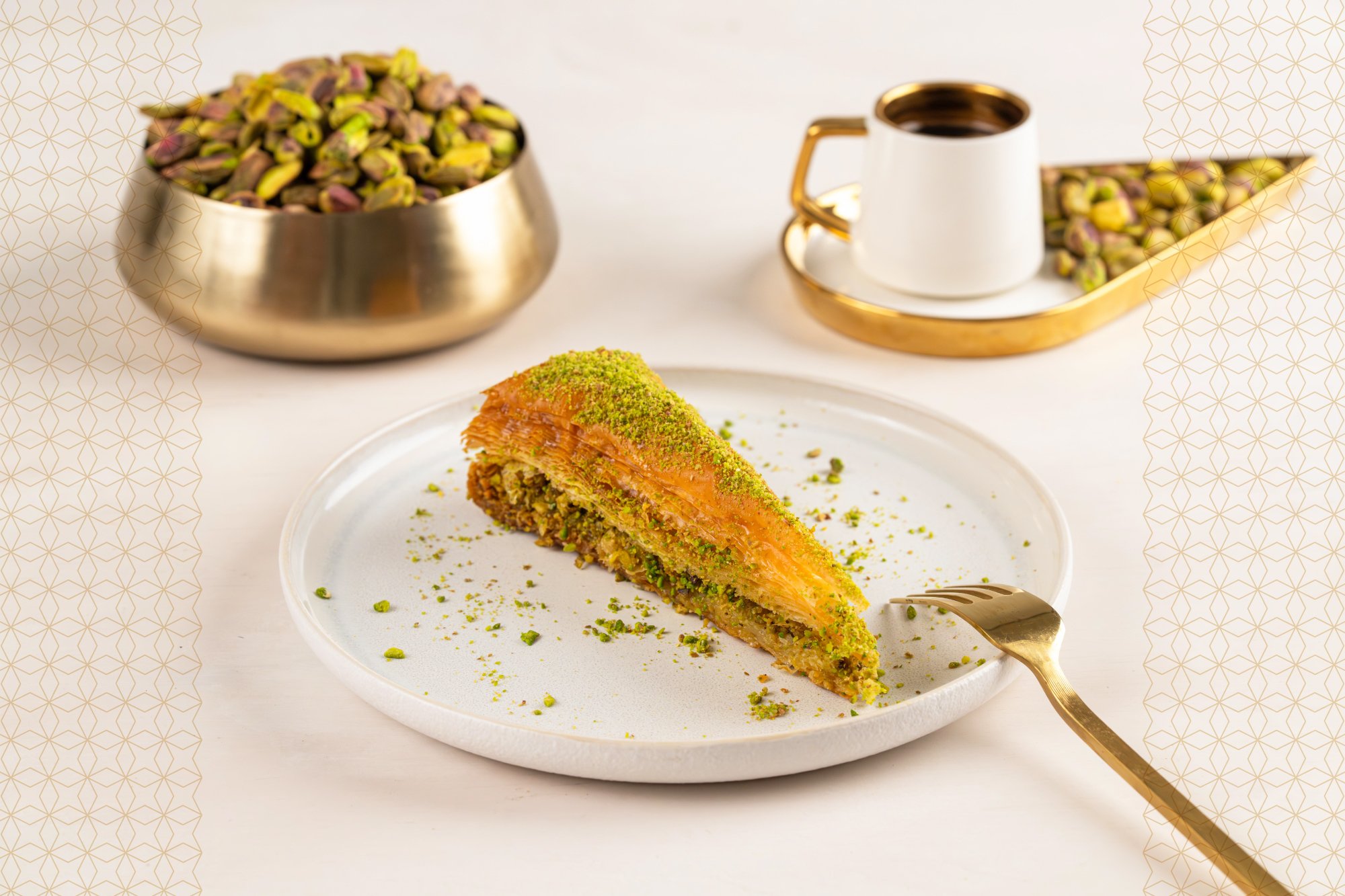Slice of pistachio baklava on a white plate, garnished with crushed pistachios, with pistachios in a brass bowl and a cup of coffee in the background.