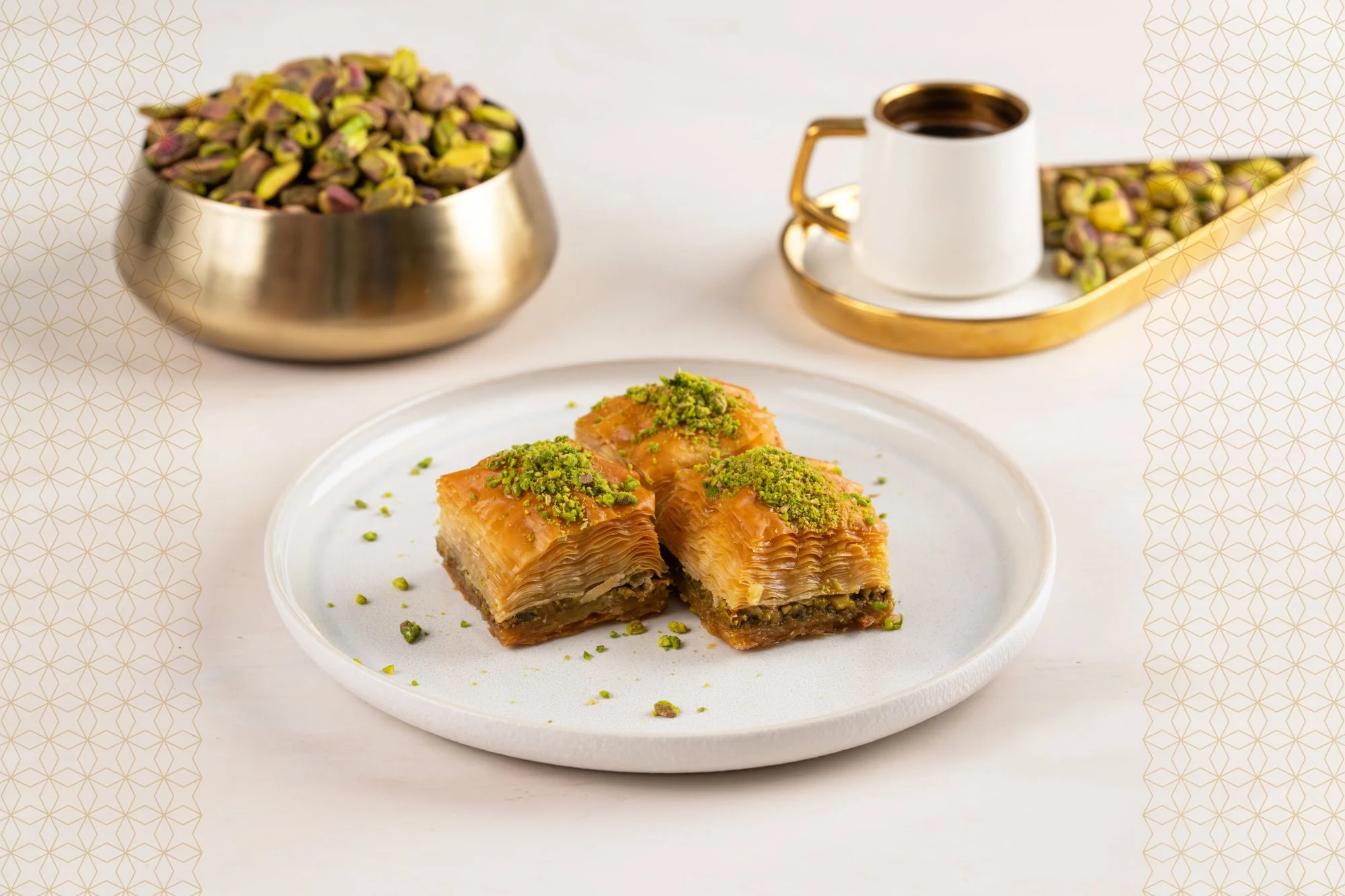 Three pieces of baklava garnished with chopped pistachios on a white plate, with a fork on the side. In the background, there is a bowl of pistachios and a cup of coffee with a saucer, all on a white table.