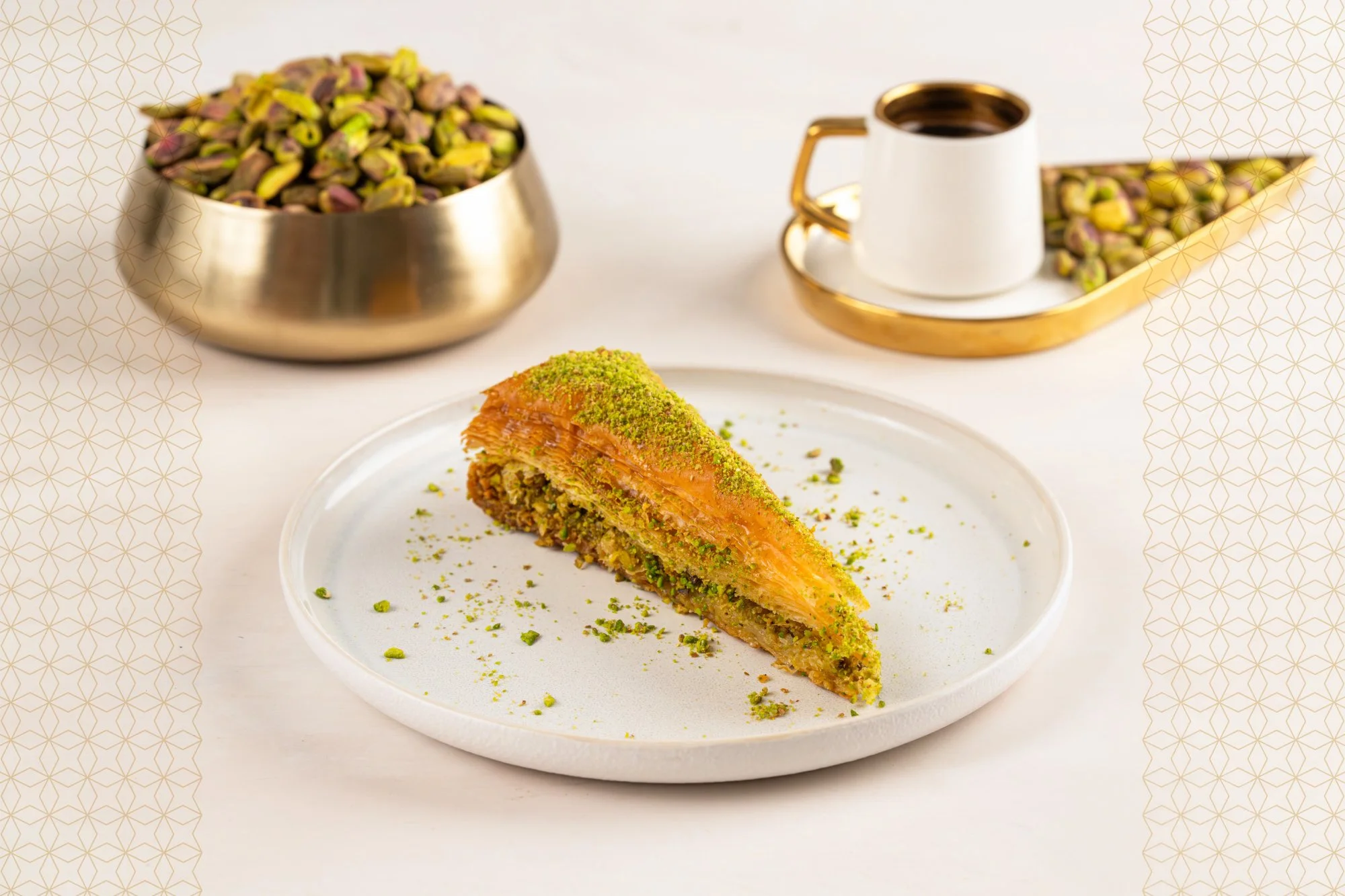 Slice of pistachio baklava on a white plate, garnished with crushed pistachios, with pistachios in a brass bowl and a cup of coffee in the background.