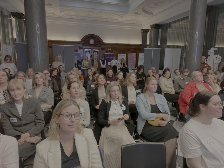 A group of professional women and men seated and listening attentively at a conference or seminar in an elegant room with columns and ornate ceiling details.