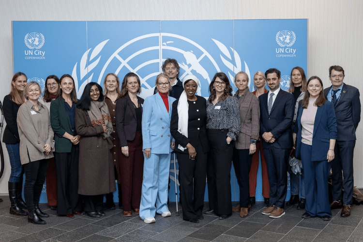 Group of diverse women and men standing together in front of a UN City Copenhagen backdrop, smiling at a conference or event.