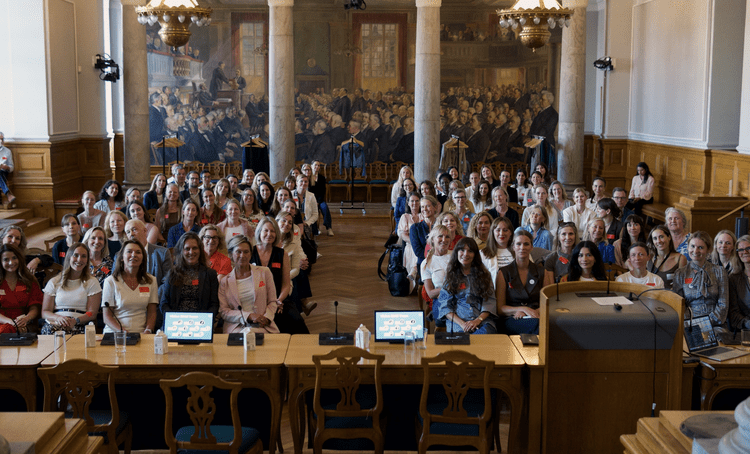 A large group of women and a few men seated in a historic hall during a conference or event. The hall features large murals, marble columns, and wooden furnishings, with a podium at the front.