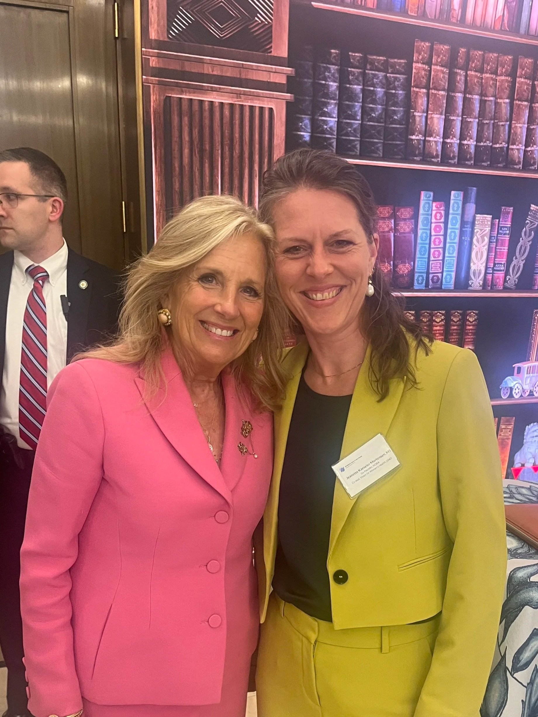 A woman with blonde hair speaking at a podium during a public event, with blurred colorful lights in the background.