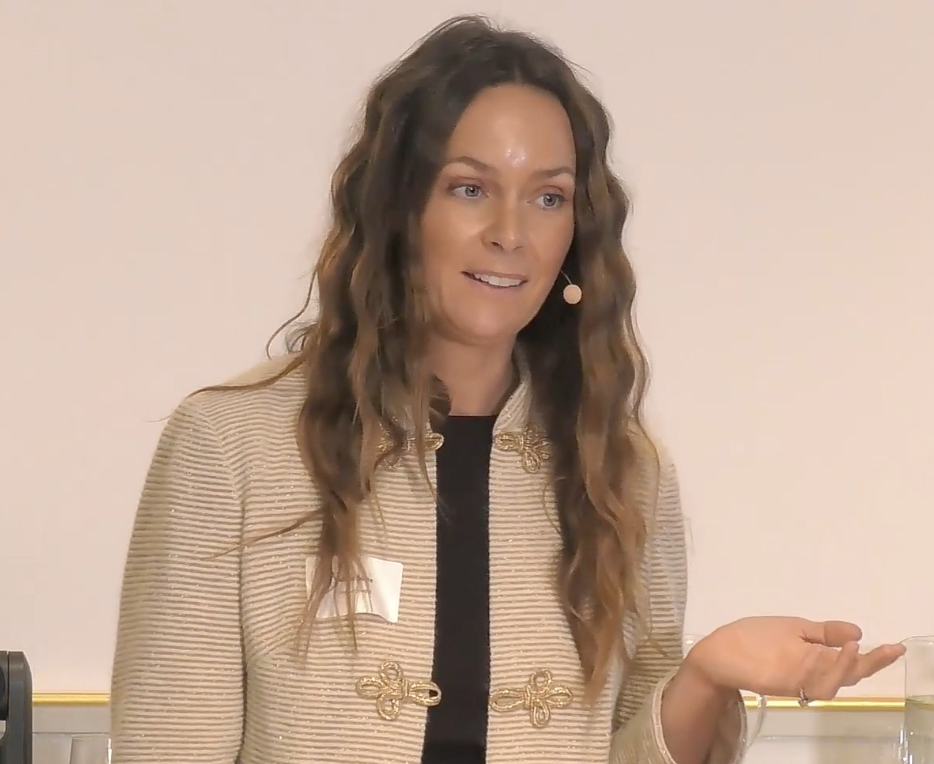 A woman is giving a presentation in a well-decorated room with a large screen displaying the word 'DO' and a glass of water on a small table.
