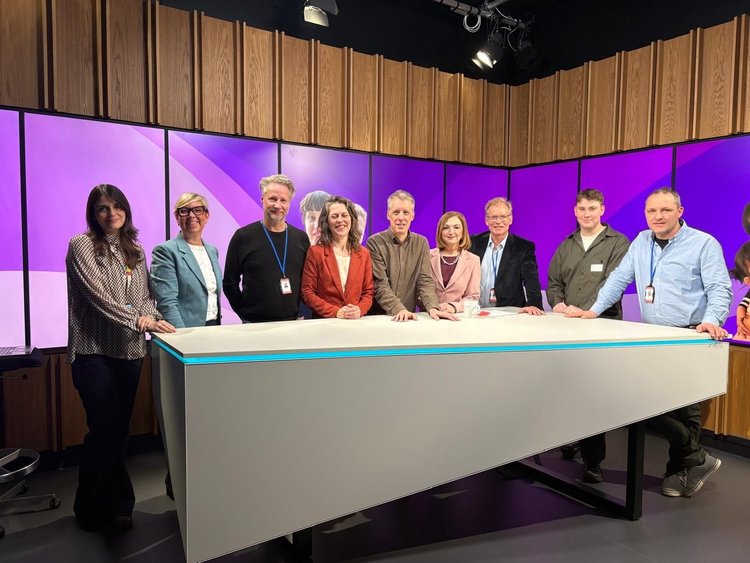 Group of nine people standing behind a conference table on a TV set with purple screens and wooden panel wall background.