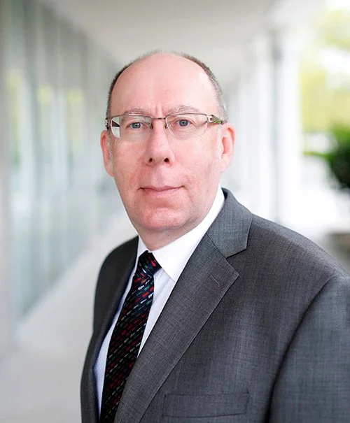 Portrait of a middle-aged man wearing glasses, a white shirt, a dark tie with red and white patterns, and a gray suit, standing outdoors near a modern building with glass windows.
