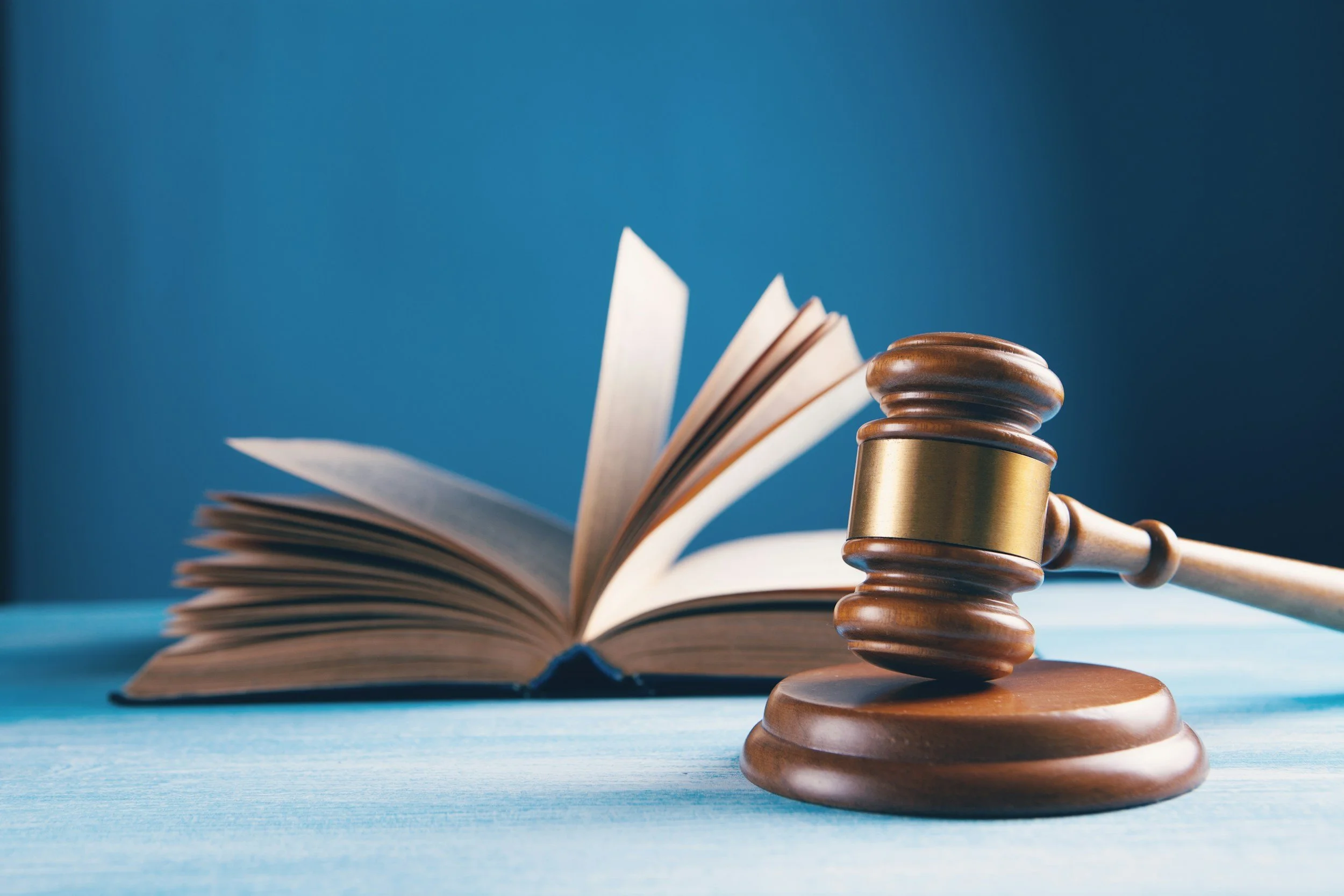 A judge's gavel resting on a round sound block in front of an open book with pages fanned out, against a blue background.