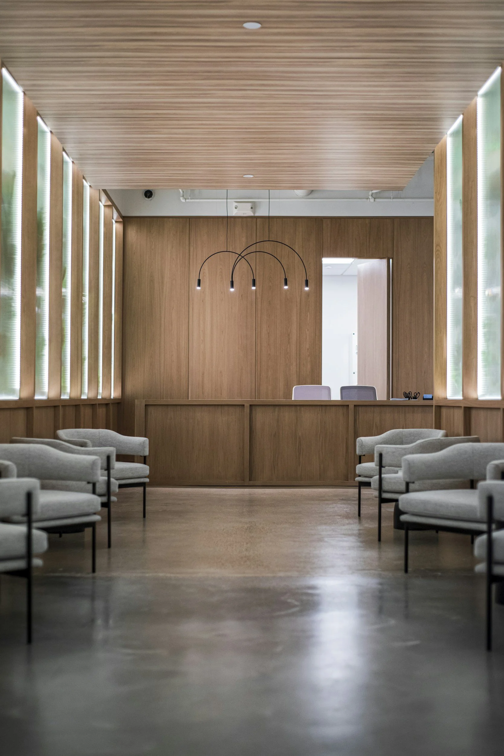 A modern, minimalist interior space with wooden panels on the walls and ceiling, white chairs arranged in rows, and a central partition. Hanging pendant lights with curved black arms and small lights are in the background near a doorway.