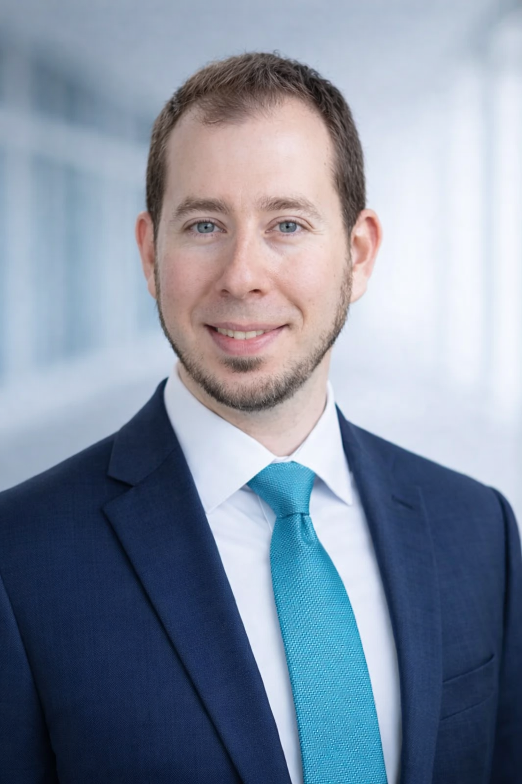 Professional headshot of a man with light brown hair, blue eyes, and light facial hair, wearing a navy blue suit, white shirt, and a turquoise tie, standing in front of a blurred modern office background.
