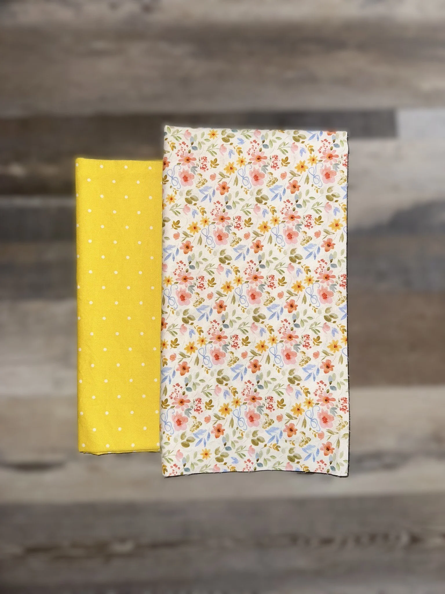 Two folded cloth napkins, one yellow with white polka dots, and one white with a floral pattern of small pink, orange, yellow, and blue flowers with green leaves.