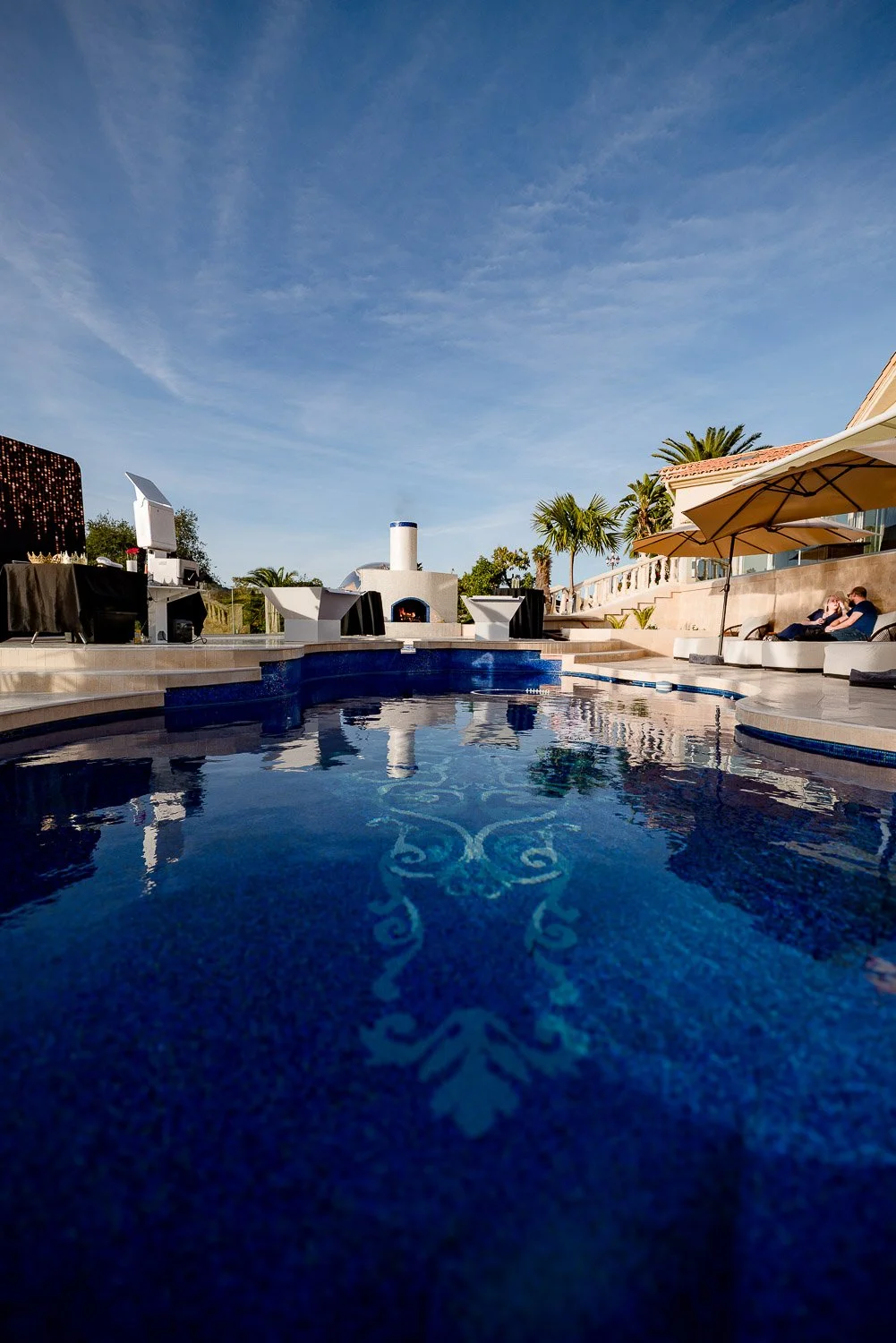 View of a luxurious swimming pool at a backyard party with palm trees, outdoor seating, umbrellas, and a clear blue sky.