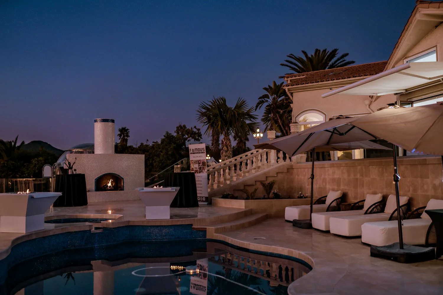 Nighttime view of a luxurious outdoor patio with a pool, fire pit, umbrellas, and lounge chairs near a house with palm trees and a fireplace.