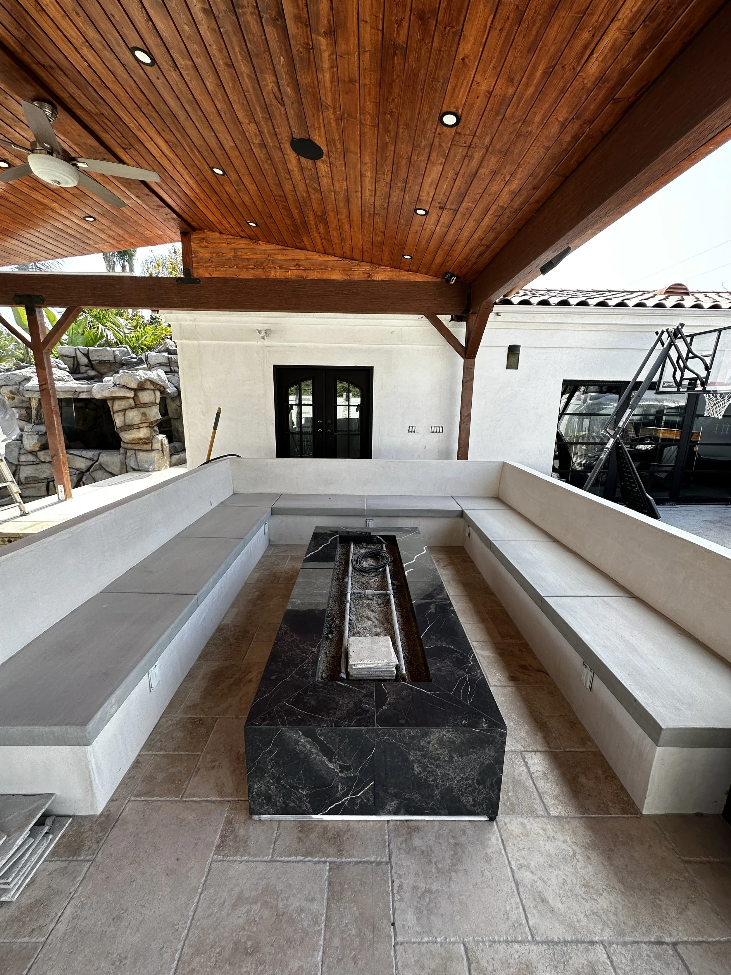 Outdoor covered patio with built-in L-shaped white concrete seating and black marble fire pit in the center, ceiling with wood paneling and recessed lighting, ceiling fan, and views of a white house wall, sliding glass door, and part of a basketball 