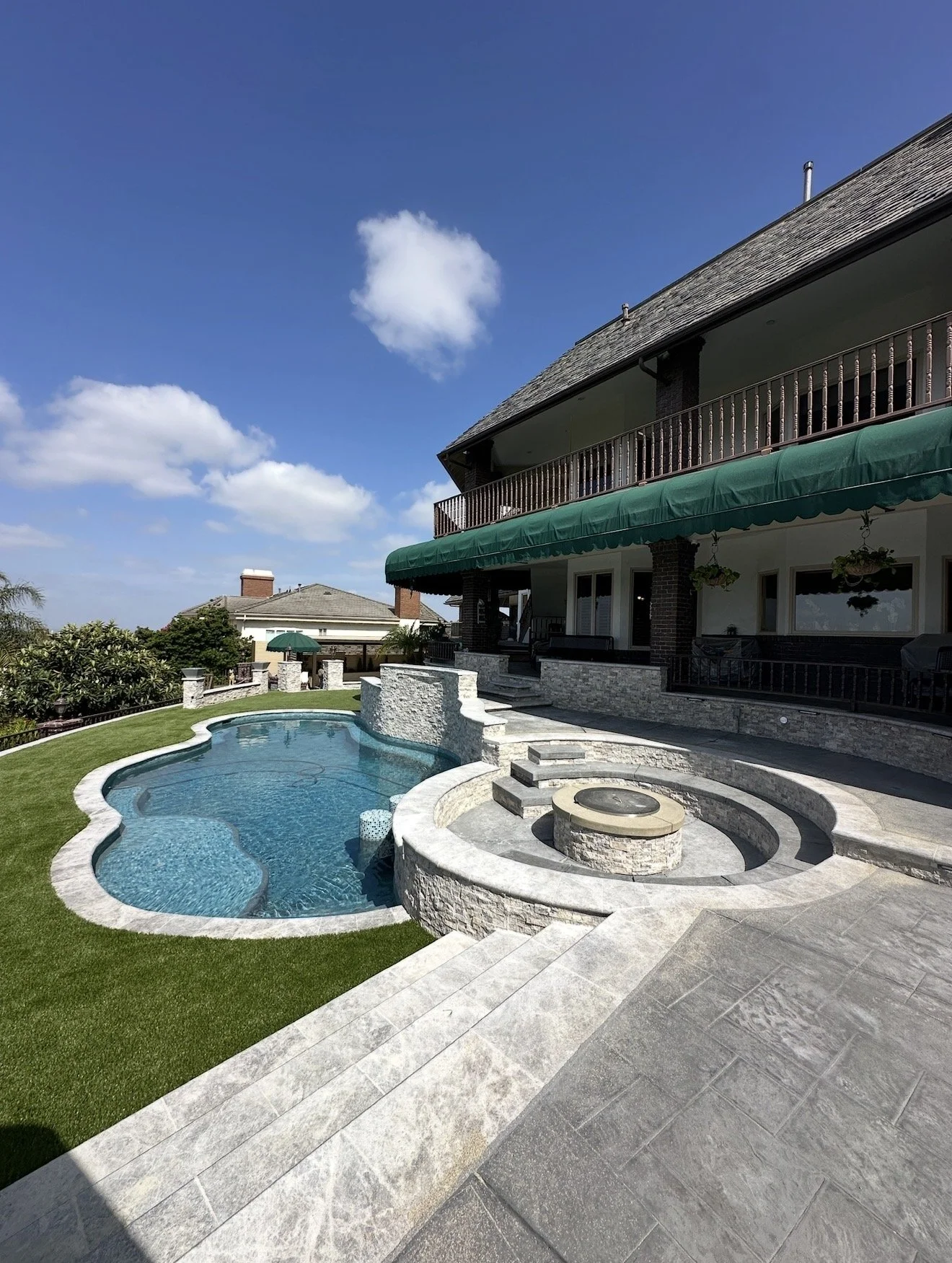 View of a backyard with a curvy swimming pool, hot tub, and patio area, adjacent to a two-story house with a covered balcony and green awning, under a partly cloudy blue sky.