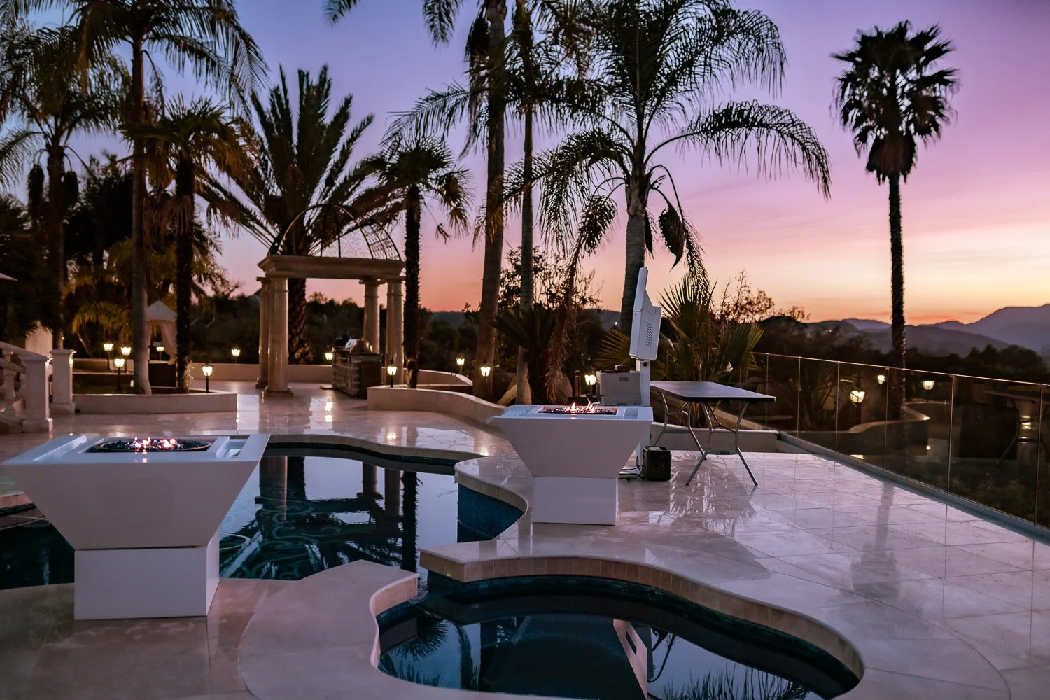 Luxury outdoor pool area with fire pits, palm trees, and mountains in the background during sunset.