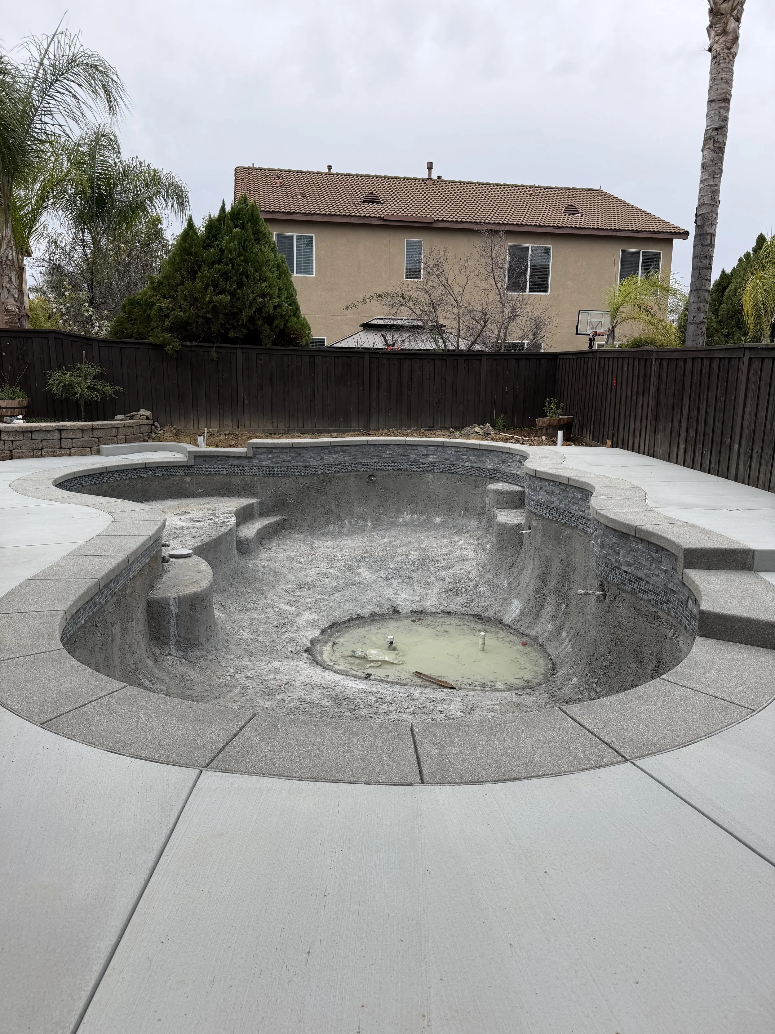 Empty swimming pool under repair with dirty water and debris, surrounded by a concrete deck in a backyard with trees and a house in the background.