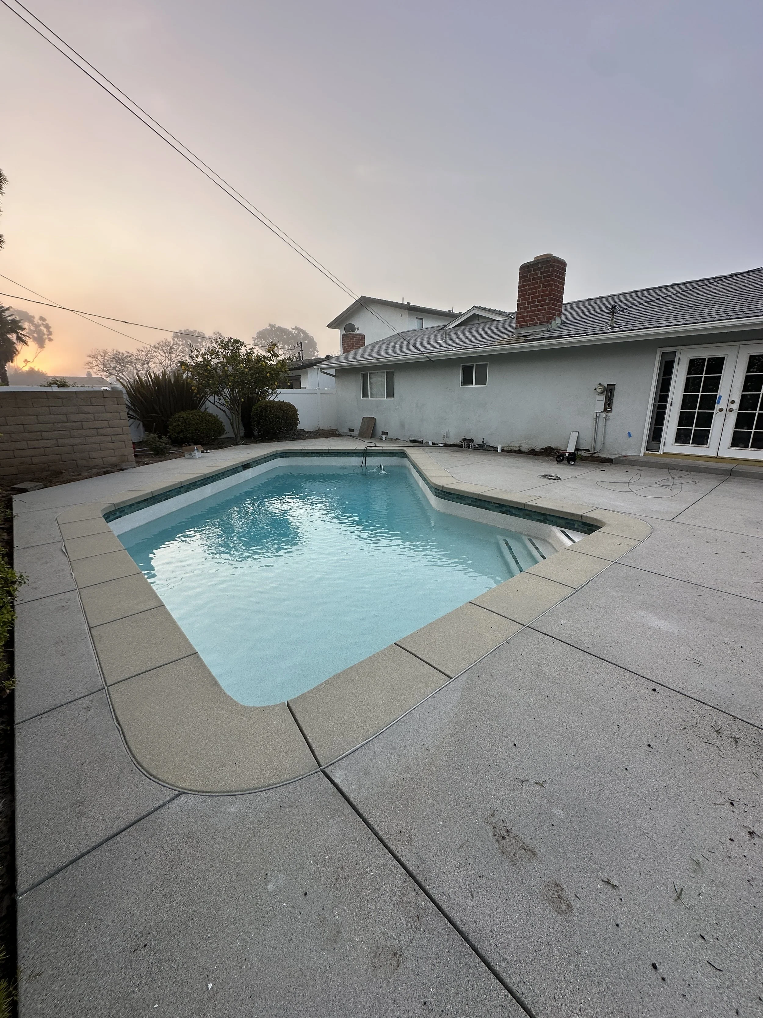 A backyard with a small swimming pool surrounded by a concrete deck, with a white house and outdoor patio doors in the background during dusk.