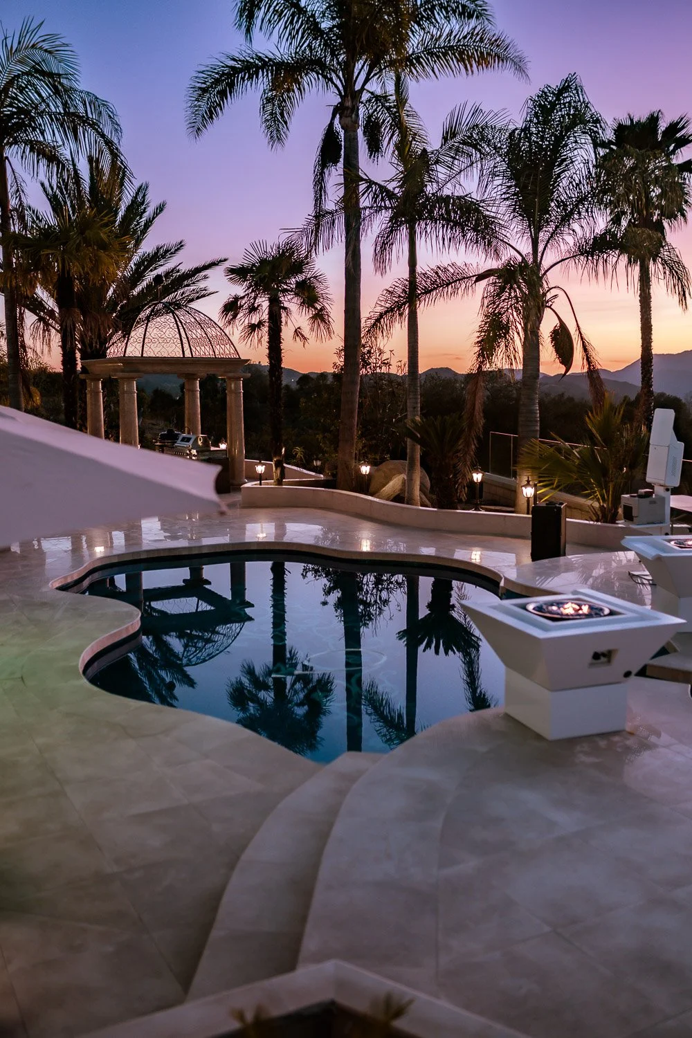 A luxurious outdoor pool area at sunset with palm trees, an open sky with purple and orange hues, and styled lounge chairs, creating a tropical resort ambiance.
