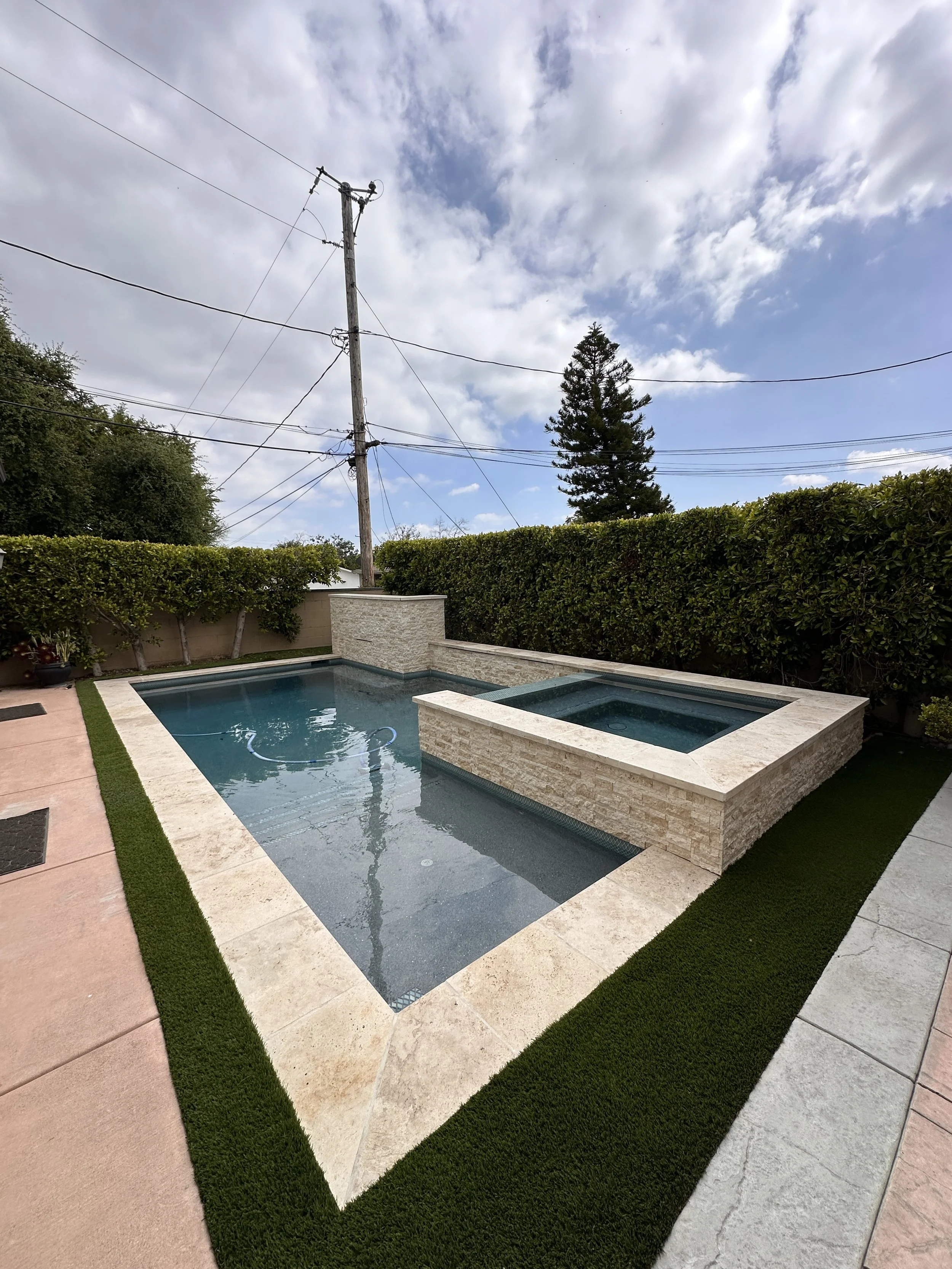 Backyard pool with built-in hot tub, surrounded by a stone border, green artificial grass, and a tall hedge. Overcast sky with clouds and a utility pole in the background.