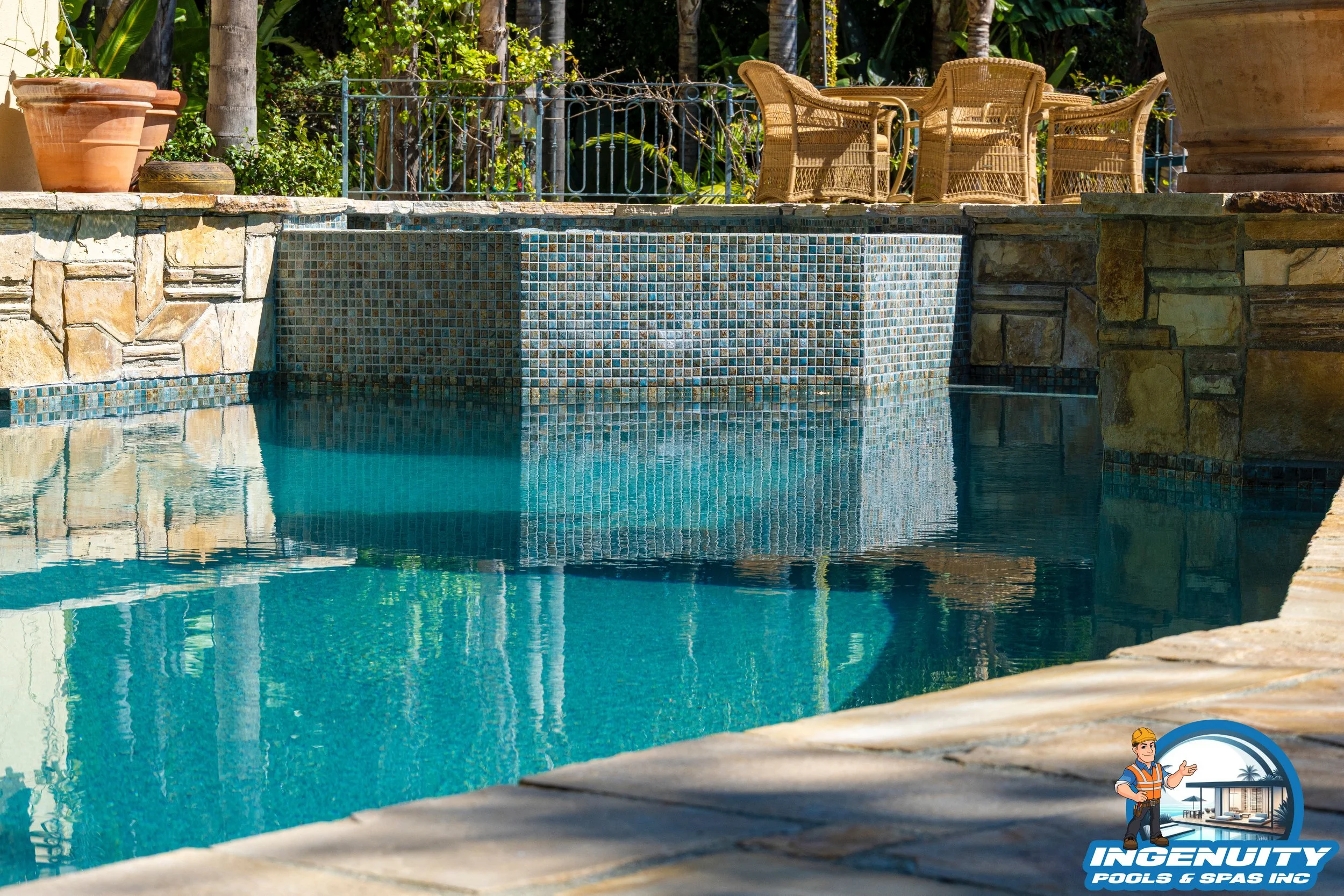 A backyard swimming pool with stone and tile walls, outdoor wicker chairs and potted plants, surrounded by greenery.
