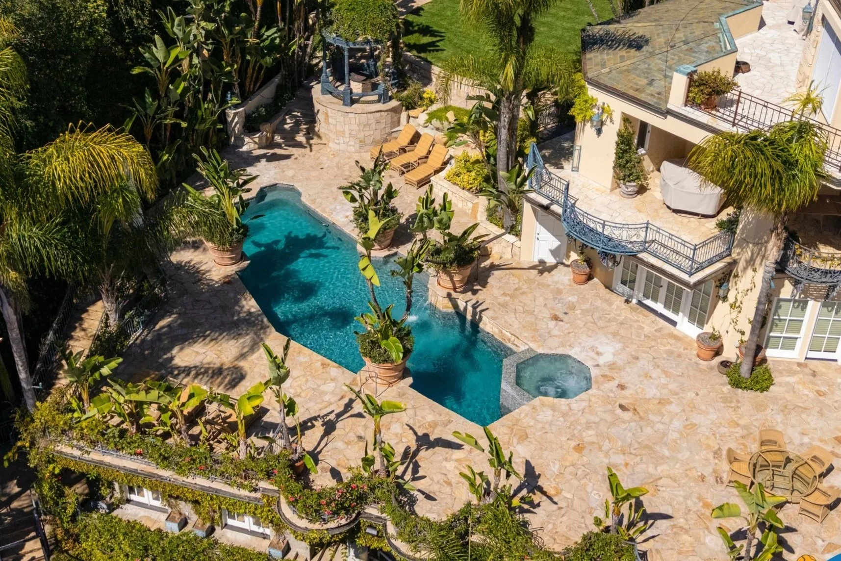 Aerial view of a luxurious backyard with a swimming pool, hot tub, and extensive stone patio surrounded by lush green plants and trees. There are lounge chairs near the pool and potted plants along the perimeter of the patio. The house has multiple b