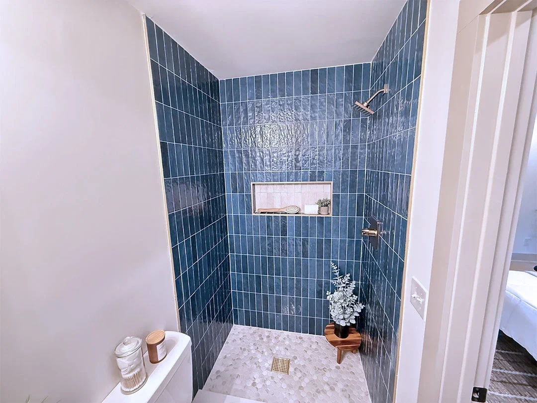Shower area with dark blue tiled walls, a small built-in shelf holding a scrub brush, white candle, and plant, a corner planter with a plant on a small wooden stand, and a drain on the pebbled floor.