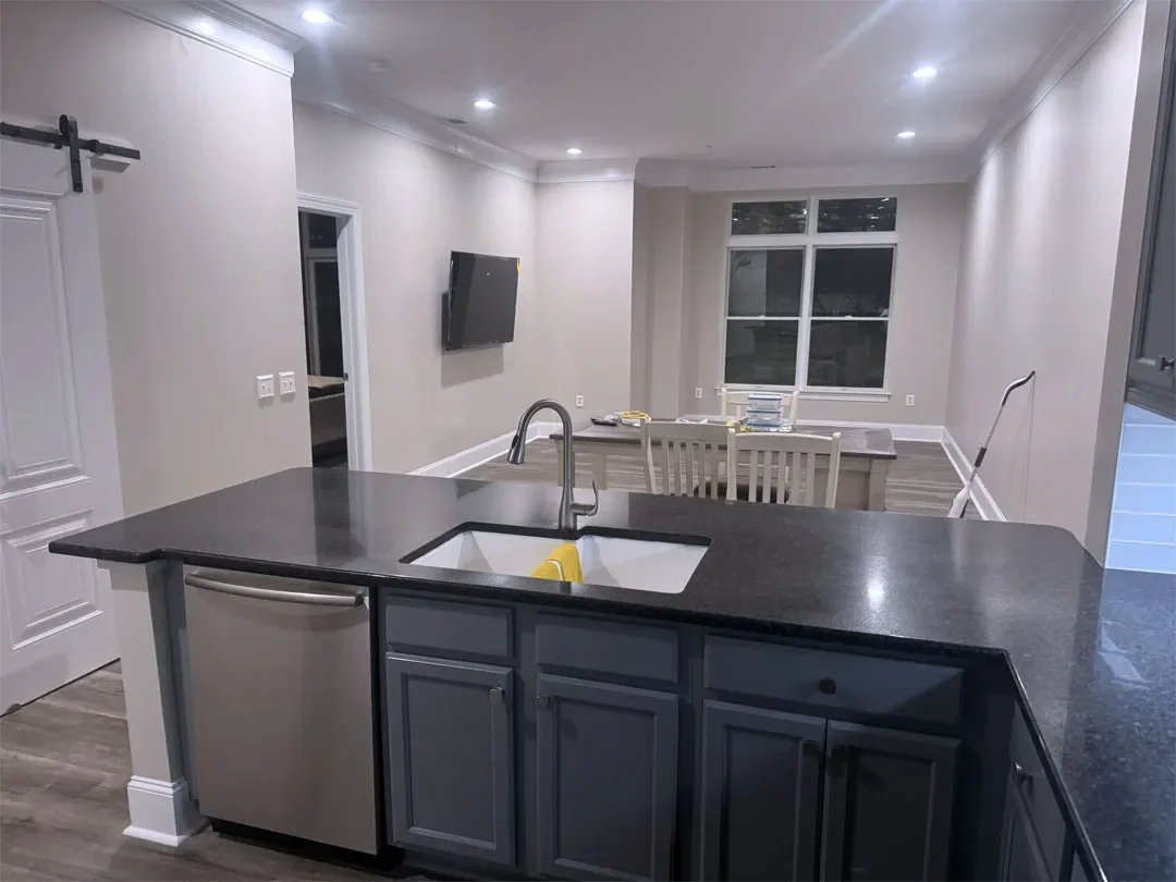 A modern kitchen with a dark countertop, a sink, and gray cabinets. In the background is a dining table with chairs and a mounted TV on the wall. Large windows with blinds are visible in the dining area.