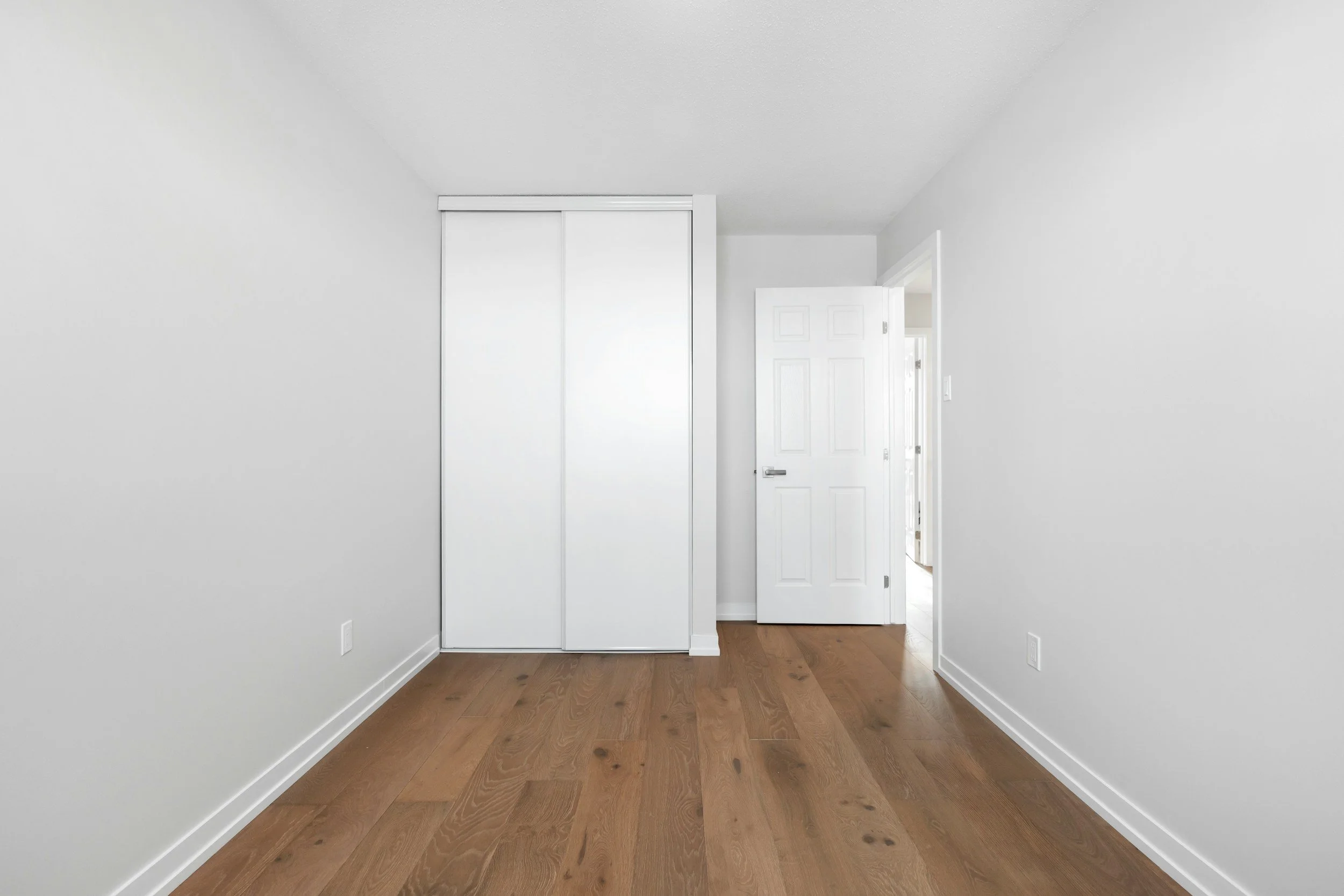 Interior view of a room with white walls, wooden flooring, a closed white closet door, and an open white door leading to another room.