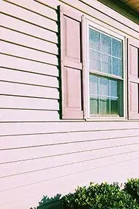 Close-up of a house exterior showing a window with purple shutters and beige siding with green bushes below.